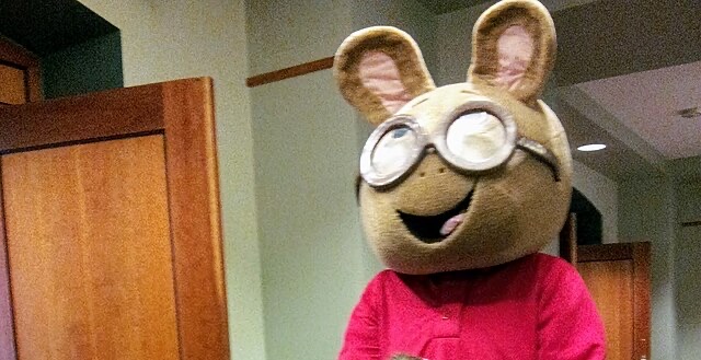 The image features Arthur Read, the beloved aardvark from the animated series "Arthur." He stands cheerfully, wearing a red sweater and a large costume head with oversized glasses that enhance his friendly demeanor. Arthur has a playful smile, embodying the character's curious and kind nature.
In the background, we see wooden paneling and a hint of a well-lit indoor space, likely a community center or event venue, which adds to the wholesome, inviting atmosphere typical of Arthur's adventures. The setting suggests a fun gathering, possibly a children’s event, where Arthur’s character would bring joy and laughter.
Humorously, Arthur's giant head contrasts sharply with the mundane background, making him look like a whimsical figure lost in a world of adults, ready to share life lessons or lead a sing-along. Overall, the image captures the essence of childhood innocence and the joy of community.