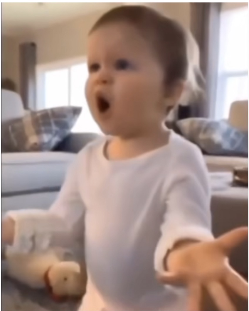 The image features a baby with a dramatically surprised expression, mouth agape as if in an exaggerated shout. The baby is wearing a white outfit and appears animated, with arms outstretched, enhancing the comical nature of the scene. The setting is a cozy living room, with soft lighting and a few pieces of furniture visible in the background, including a couch adorned with plaid cushions. 

This scene captures a moment of pure shock or disbelief, making it relatable and humorous, often used in memes to express exaggerated reactions. The baby's demeanor, full of innocence yet intense surprise, brings a lighthearted touch to the image, as if it's reacting to some unexpected news. The overall atmosphere is warm and inviting, framing the baby’s expression in a comfortable domestic setting, emphasizing the relatability of the moment.