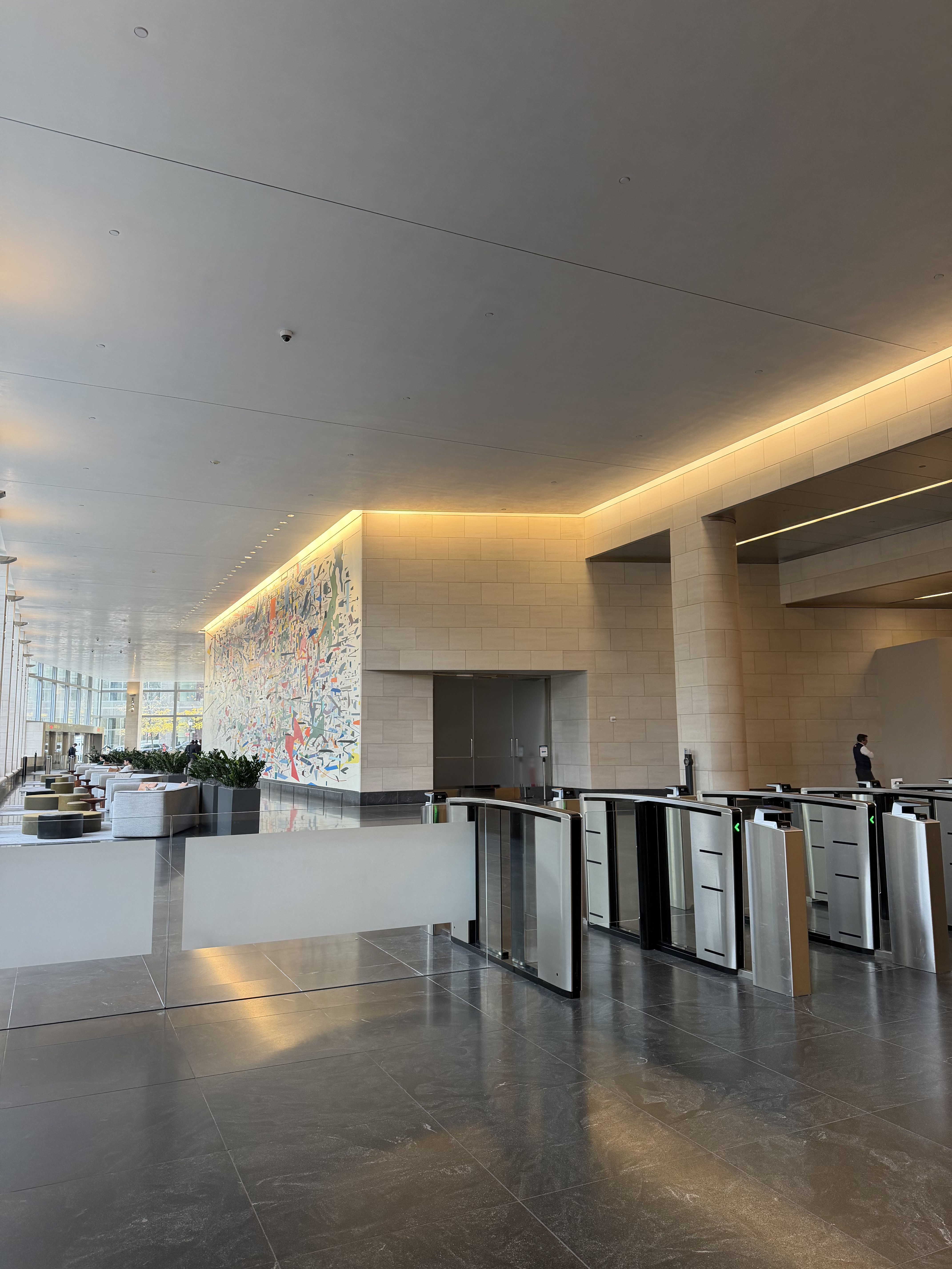 This image presents a vast, modern corporate lobby, exuding a sleek, almost futuristic professionalism. A lone man in a dark vest stands near a row of gleaming security turnstiles, appearing calm and observant, perhaps contemplating the existential dread of another workday. The turnstiles, with their glowing green arrows, seem to patiently await their next human cargo, silently judging each swipe. To the left, a comfortable seating area invites weary souls, while a vibrant, abstract mural splashes color across a distant wall, a playful rebellion against the muted tones. Large windows reveal an autumnal cityscape, hinting at the world outside this controlled environment. The polished dark floor reflects the warm strip lighting, adding to the sophisticated, yet slightly sterile, atmosphere. No famous content is recognized.