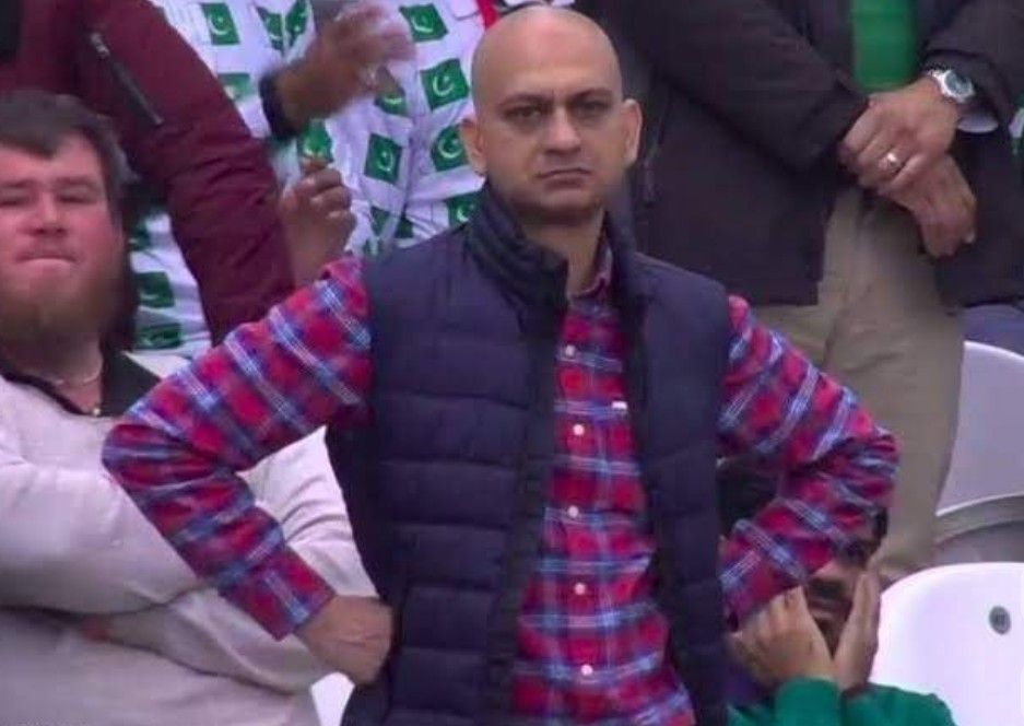 The image captures the widely recognized 'Disappointed Fan' meme, featuring a bald man with a profoundly exasperated and stern expression, hands firmly on his hips. He is dressed in a vibrant red and blue plaid shirt beneath a dark blue puffer vest. The background clearly shows a crowd, with several spectators wearing white shirts emblazoned with the green crescent and star of the Pakistani flag, indicating a cricket match, likely from the 2019 Cricket World Cup. His intensely relatable demeanor, embodying universal disappointment or frustration, is the core humorous element, making this image a popular reaction meme for countless situations beyond sports.