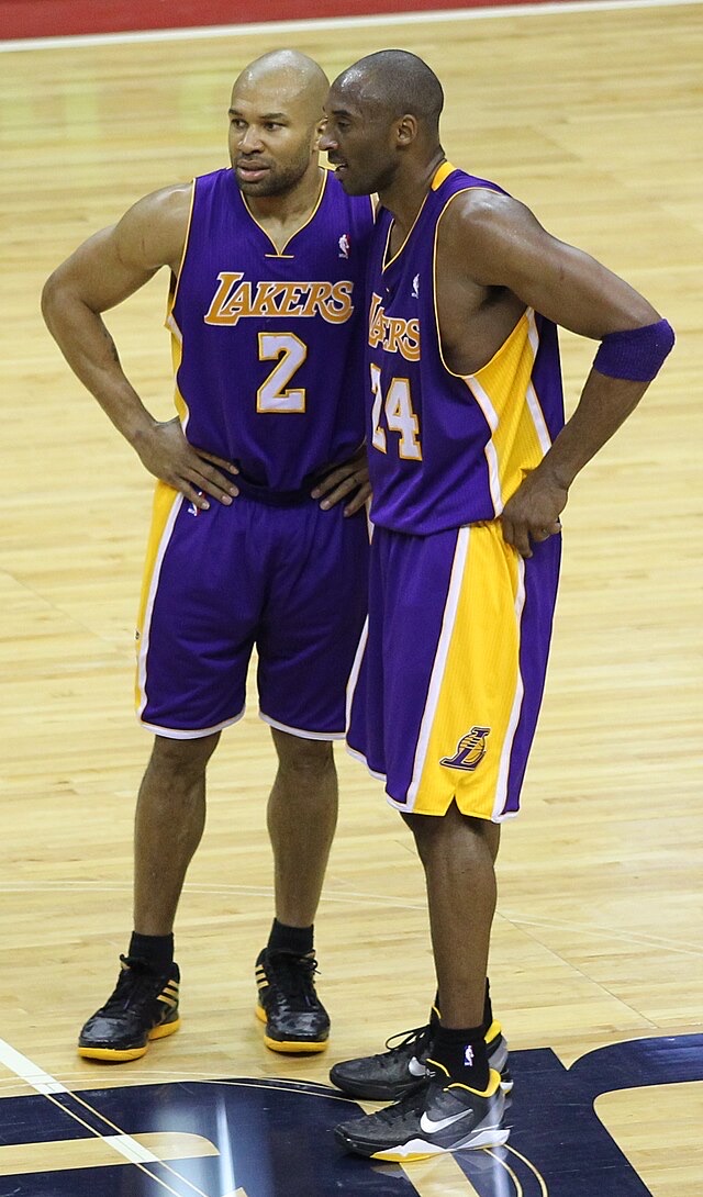 This image captures NBA legends Kobe Bryant (#24) and Derek Fisher (#2) of the Los Angeles Lakers, standing on a polished wooden basketball court. Both players, bald and sporting their iconic purple and gold uniforms, have their hands on their hips, displaying a focused yet relaxed demeanor, perhaps discussing strategy or a recent play. Fisher, on the left, wears black and yellow Adidas sneakers, while Kobe, on the right, sports black and yellow Nike shoes, a subtle brand rivalry on the same team. The court's markings and the visible NBA logo confirm the professional basketball setting. It's a classic moment of two teammates, seemingly deep in thought, perhaps wondering if they remembered to turn off the stove before the game.