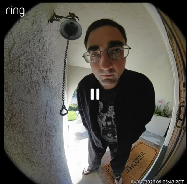 In this image, we see a young man peering closely into a doorway camera, his demeanor is curious yet slightly apprehensive. He wears a casual outfit consisting of a black cardigan, a graphic t-shirt, and loose pants, along with flip-flops, which adds an element of relaxed informality. His large glasses magnify his eyes, giving him a slightly comical expression as he leans in, possibly trying to see who is on the other side of the door.
The background features a simple porch area with a welcome mat, hinting at a homey setting. A small flower pot adds a touch of color and life to the scene. The image could evoke a humorous vibe, as the man's exaggerated posture and intense gaze resemble a scene from a sitcom, where a character is caught in a funny, awkward moment of trying to sneak a peek.