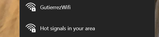 This image displays a digital interface showcasing two Wi-Fi networks, each with a distinct 'personality.' On a dark grey background, 'GutierrezWifi' appears as a reliable, no-nonsense network, simply presenting itself. Below it, 'Hot signals in your area' exudes a playful, almost flirtatious demeanor, winking at potential users with its cheeky name, promising both strong connectivity and a hint of intrigue. Both networks are secured, indicated by the padlock icons, suggesting they are confident in their offerings but not easily accessible. The blurry, warm-toned background on the left, possibly wood, subtly grounds these digital entities in a physical, perhaps cozy, environment, adding a touch of real-world context to their virtual broadcast. The humor lies in the 'Hot signals' network, anthropomorphized as a confident, attention-grabbing character in the digital neighborhood.