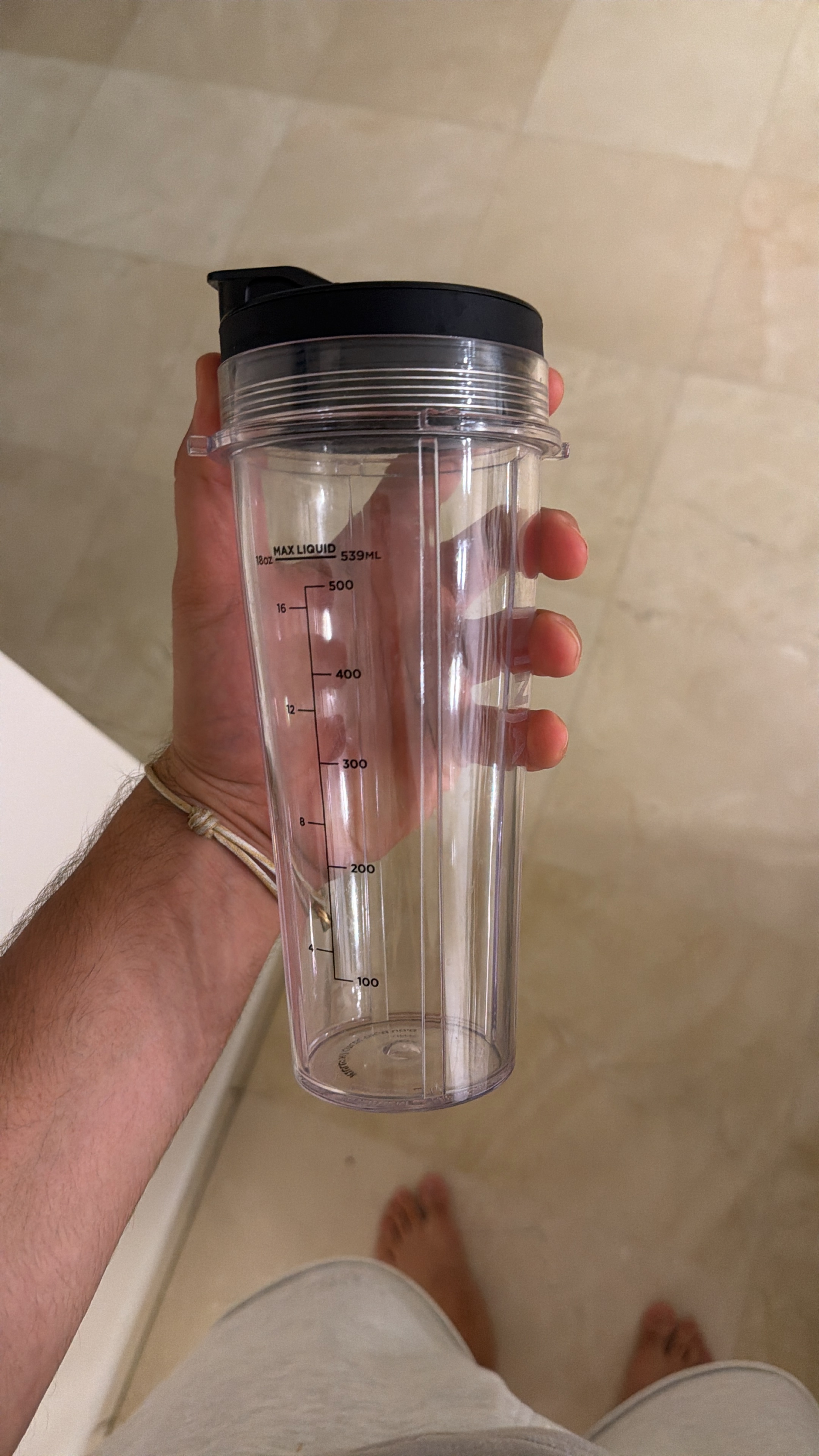 The image prominently features a clear plastic blender cup, complete with a black lid and precise measurement markings up to 539ml, held firmly by a person's hand. The cup, with its utilitarian design, appears ready for its next liquid adventure, perhaps a smoothie or protein shake. The hand, showing some hair and a simple light-colored string bracelet, suggests a casual, at-home environment. In the background, the person's bare feet and the edge of light-colored clothing are visible on a neutral-toned tiled floor, reinforcing a relaxed, domestic setting. The humor lies in the cup's earnest, measured presence against the candid, almost accidental inclusion of the person's feet, as if the cup is the main character in a very important, yet utterly ordinary, daily ritual. No famous content is present.