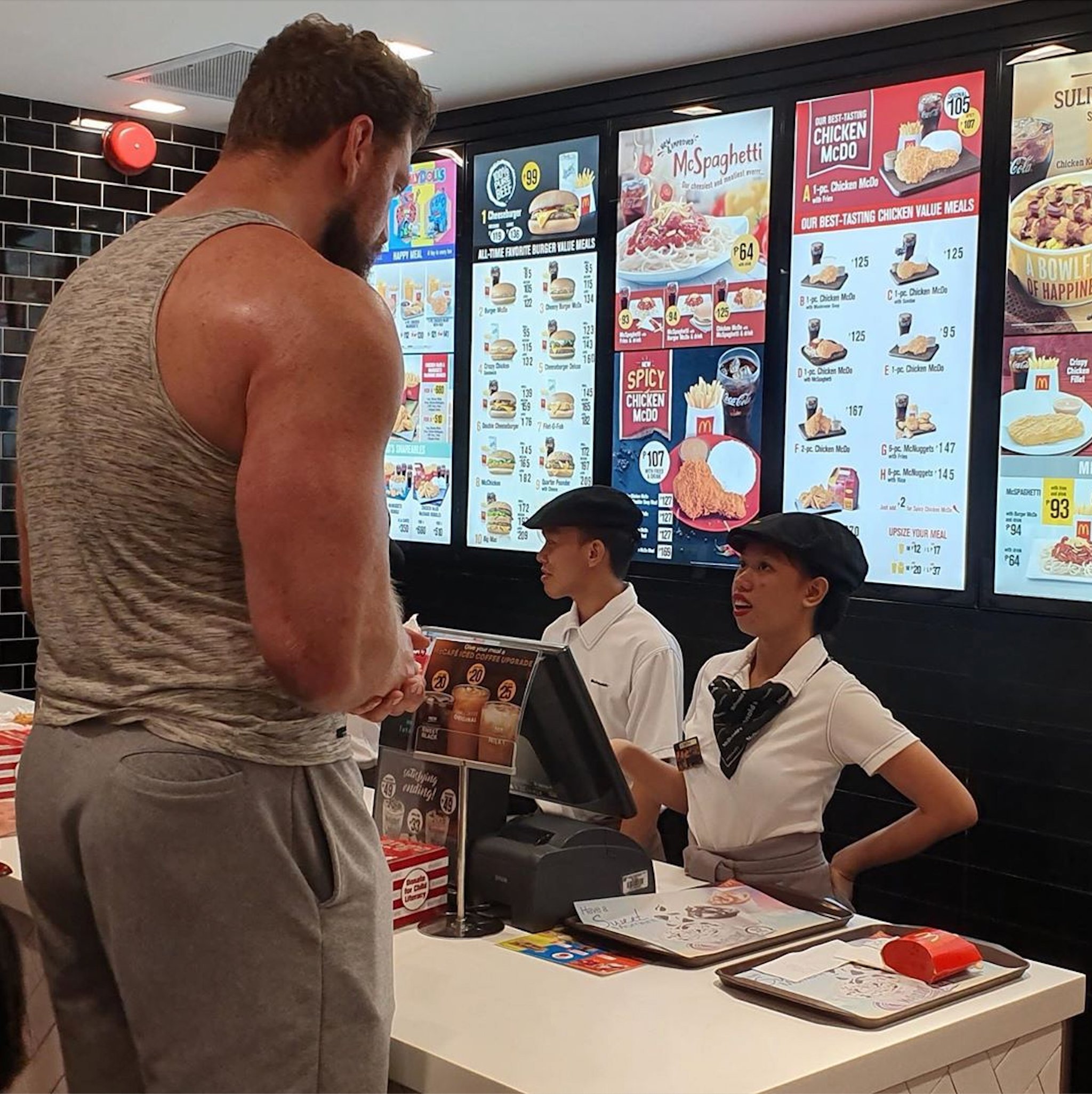 A towering, muscular man in a grey tank top and sweatpants leans over a fast-food counter, his gaze directed downwards. His imposing physique contrasts humorously with the setting. Behind the counter, a female employee in a white uniform and black cap looks up at him with a subtly amused or surprised expression, likely due to their significant height difference. Beside her, a male colleague appears more stoic. The background features brightly lit menu boards displaying various meal options, including "McSpaghetti" and "Chicken McDo," indicating a regional fast-food establishment. The scene captures a common, yet comically exaggerated, interaction at a busy eatery, where the customer's sheer size creates a memorable moment for the staff.