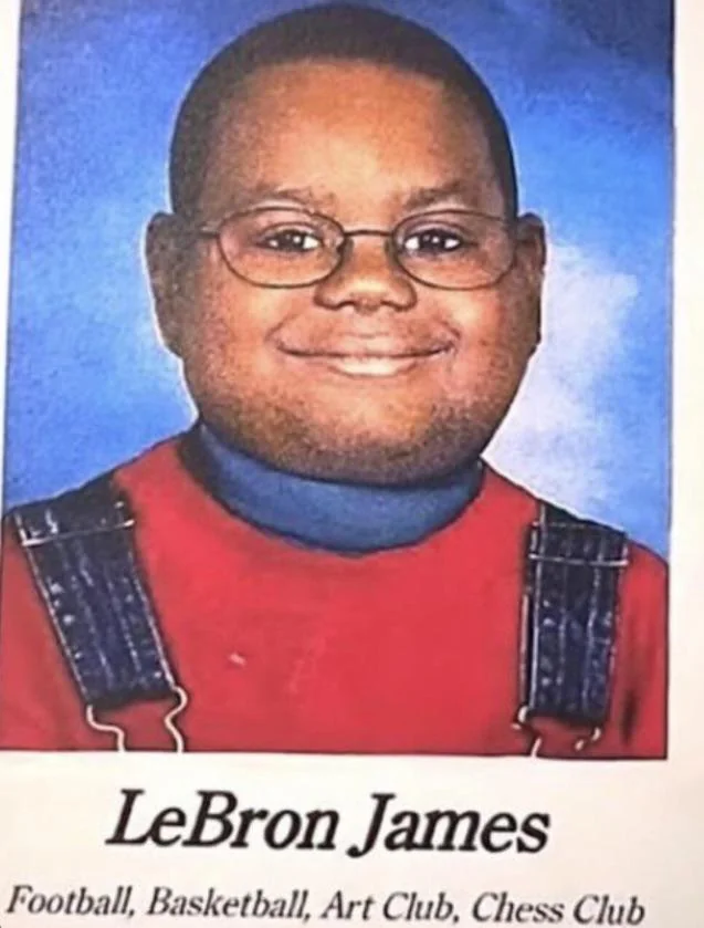 This image is a humorous meme, a yearbook-style portrait of a young boy identified as 'LeBron James.' The child, with dark skin and round glasses, sports a wide, slightly awkward but endearing smile, looking directly at the camera. He wears a blue turtleneck under bright red overalls, a classic childhood ensemble. The plain blue background is characteristic of school photos. The humor arises from the stark contrast between this innocent-looking child and the globally recognized basketball superstar. The listed activities, 'Football, Basketball, Art Club, Chess Club,' playfully suggest a surprisingly diverse range of interests for someone who would become an athletic icon, amplifying the meme's comedic effect.