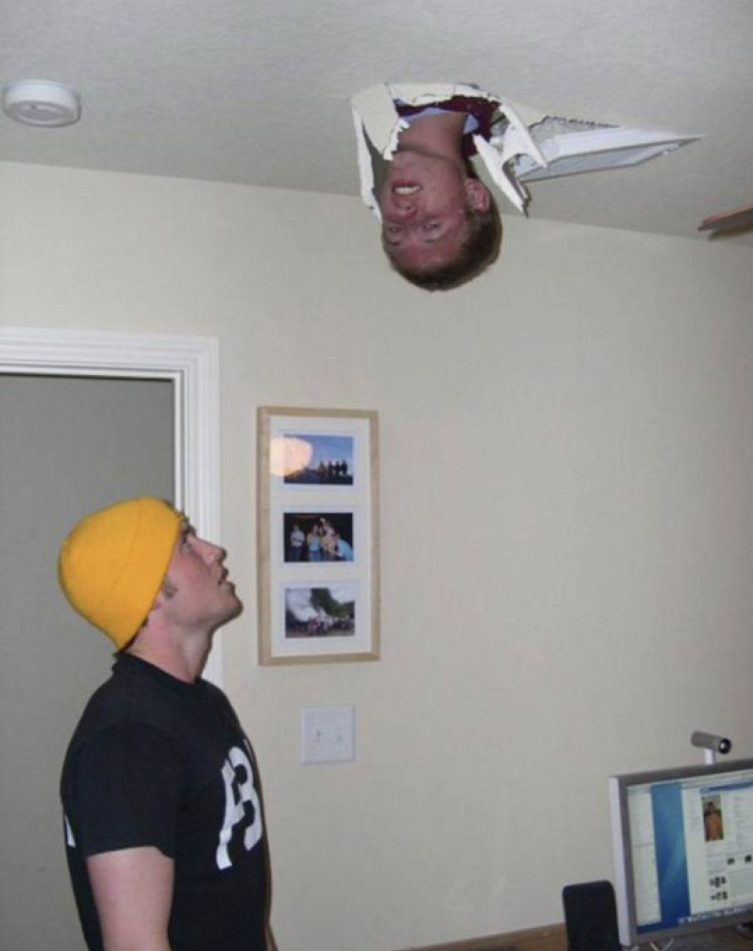 This image captures a truly bizarre and humorous domestic scene. A man's head, with a slightly dazed or amused expression, protrudes upside down from a jagged hole in the ceiling, as if he's just dropped in from another dimension. Below, another man, sporting a bright yellow beanie and a black t-shirt, gazes upwards with a look of utter bewilderment and perhaps a hint of 'what just happened?' The background is a typical room with light-colored walls, a white door frame, and a vertical frame holding three small pictures. A desk with a computer monitor and speakers is visible to the right. The plain, everyday setting starkly contrasts with the surreal sight of a head emerging from the ceiling, making the situation inherently comical. The background serves as a mundane stage for this unexpected, gravity-defying prank or mishap. I could not identify any famous or recognizable content in this image.