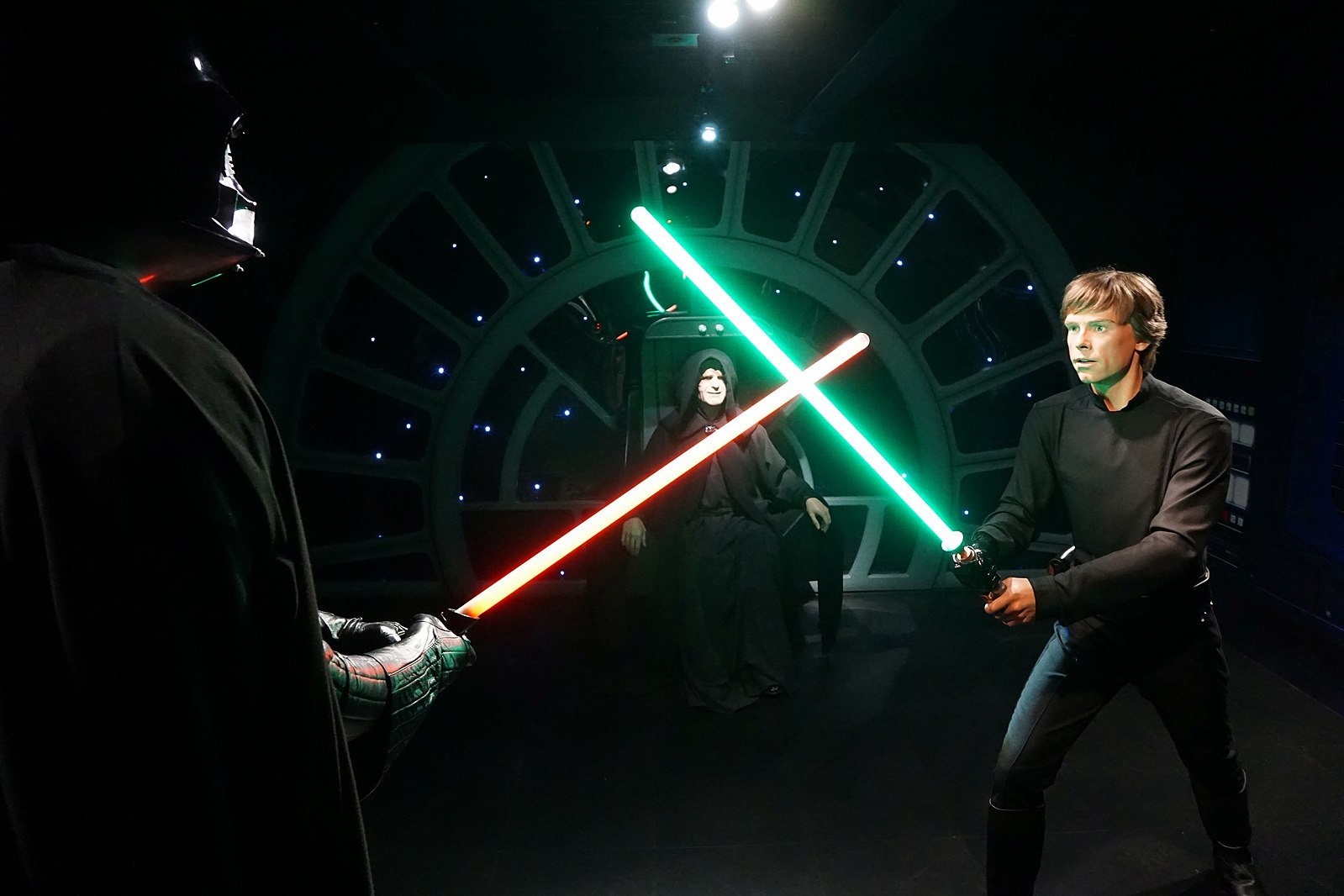 This image vividly recreates the iconic climax of Star Wars: Episode VI – Return of the Jedi, featuring wax figures of Darth Vader, Luke Skywalker, and Emperor Palpatine. Luke Skywalker, holding his green lightsaber, stands in a defensive stance, his face conveying a mix of determination and apprehension. Darth Vader, seen from behind, wields his red lightsaber, ready for the clash. Emperor Palpatine, seated on his throne in the background, observes the duel with a chillingly calm and sinister demeanor. The circular window of the Death Star Throne Room, with its distant starfield, perfectly frames this pivotal confrontation. The humor arises from these intensely dramatic characters being frozen in time as wax figures, forever locked in their epic, yet utterly static, struggle.