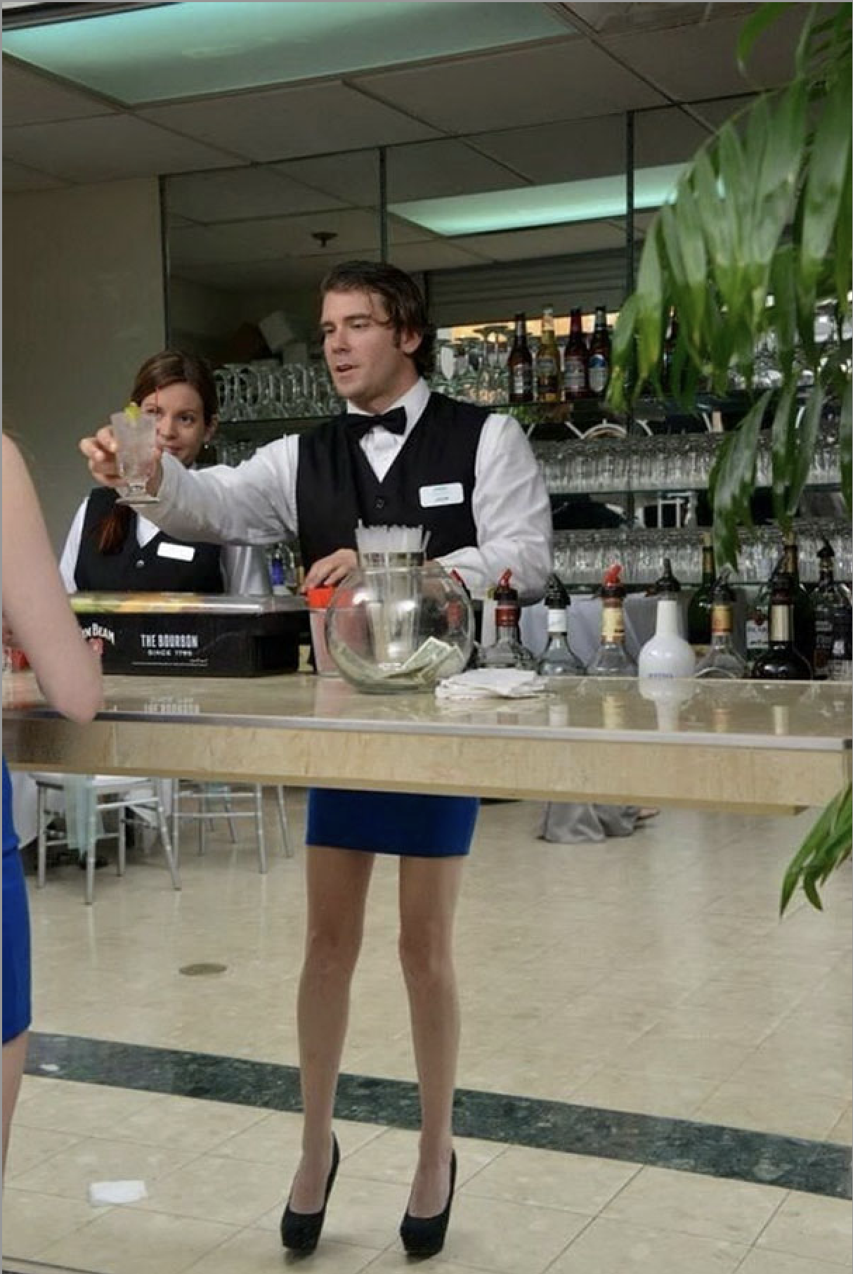 A male bartender, dressed in a black vest and bow tie over a white shirt, humorously sports a blue mini-skirt and black high heels. He confidently extends a cocktail towards an unseen patron, a slight smile on his face, suggesting he's fully embracing his unconventional uniform. Behind him, a female colleague with a more traditional uniform looks on, while a well-stocked bar with bottles and glassware reflects in a mirrored wall. The setting is clearly an event bar, with a tip jar visible, and the background perfectly complements the foreground's service activity. The playful attire of the male bartender is the central humorous element, adding a lighthearted twist to the professional setting. I did not find any famous or recognizable content in this image including people, shows, movies, locations, brands, events, popular memes, or cultural references.