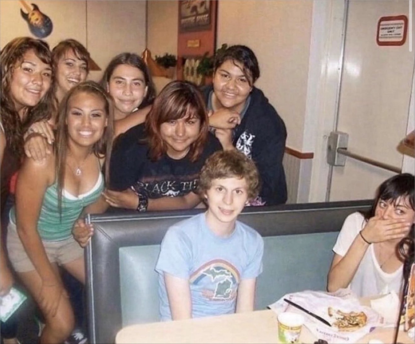 This iconic meme-worthy image captures actor Michael Cera sitting awkwardly at a booth, sporting his signature shy smile and a light blue t-shirt with a rainbow graphic. He is surrounded by a group of six smiling young women, who appear delighted to be posing with him. To his right, actress Aubrey Plaza is visible, covering her mouth, seemingly suppressing laughter or surprise, adding to the image's comedic timing. The background suggests a casual family restaurant or arcade, possibly Chuck E. Cheese, with booth seating, a guitar decoration, and an 'Emergency Exit Only' sign above Plaza. The humor stems from Cera's characteristic demeanor amidst the enthusiastic fans and Plaza's amused reaction, creating a perfectly candid and relatable celebrity encounter.