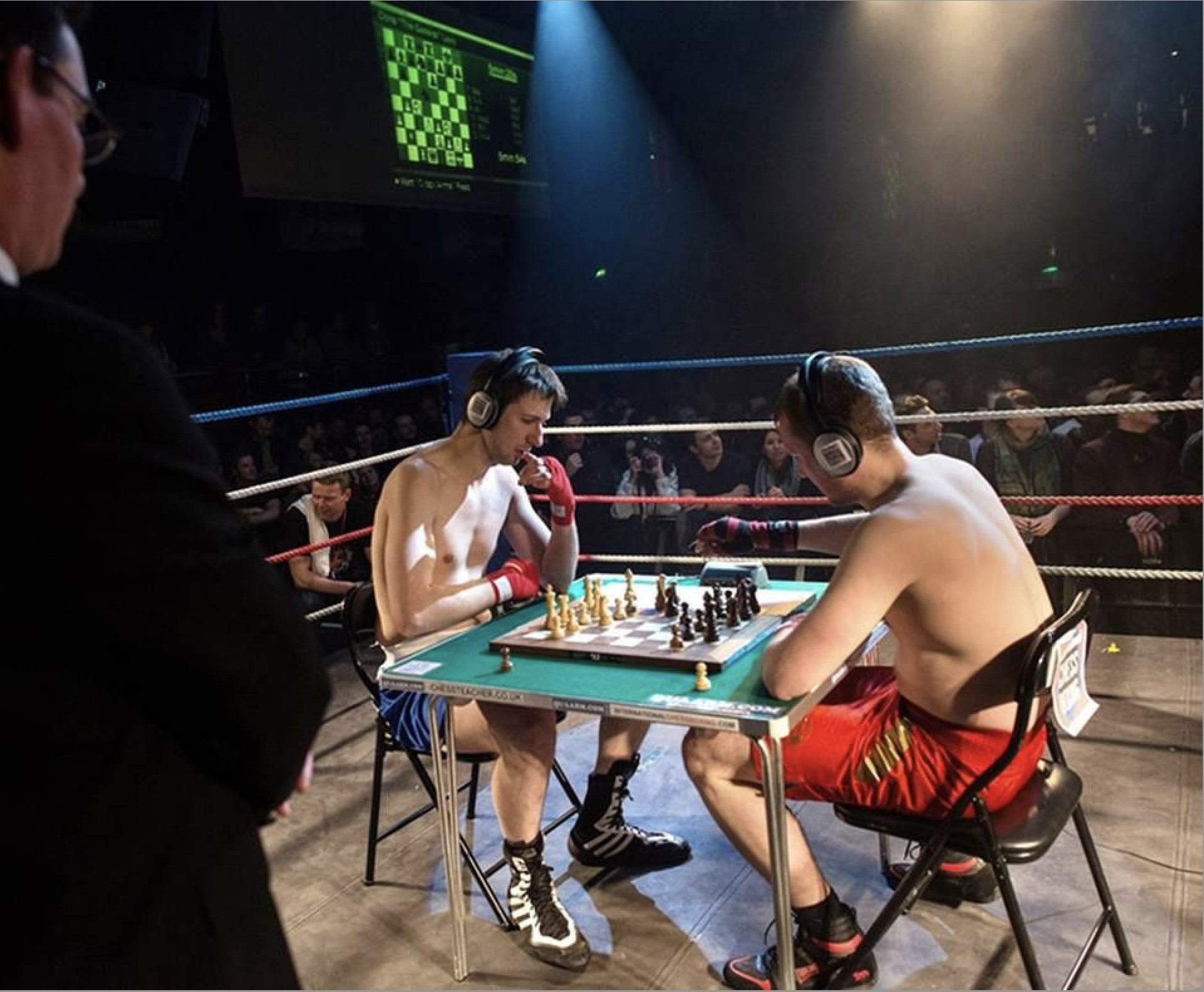 The image depicts a unique 'chess boxing' event, where two shirtless men, wearing boxing gloves and headphones, are deeply engrossed in a chess game set up on a table within a boxing ring. Both subjects exhibit intense concentration, with one player thoughtfully holding his chin, contemplating his next move. The background prominently features a boxing arena with ropes, an audience, and a large screen displaying a digital chess board, clearly establishing the context of this hybrid sport. The humor stems from the ironic juxtaposition of the quiet, intellectual game of chess with the aggressive, physical setting of a boxing match. This unusual cultural event blends mental strategy and physical prowess into a compelling, albeit quirky, spectacle.