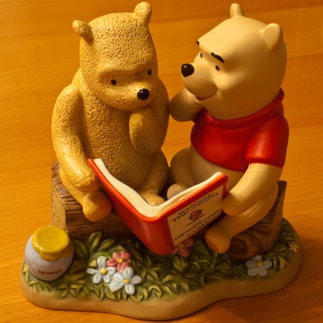 This charming figurine depicts Winnie the Pooh, in his classic red shirt, engrossed in reading "Winnie the Pooh and the Honey Tree" to his textured companion, identified as Piglet by the context. Pooh appears to be animatedly narrating, perhaps reliving his own adventures, while Piglet listens with a wonderfully pensive and curious demeanor, hand thoughtfully placed on his chin. They are seated on rustic logs amidst a patch of green grass and delicate flowers, evoking the serene Hundred Acre Wood. A classic "HUNNY" pot sits nearby, a humorous reminder of Pooh's insatiable appetite, even while he's immersed in a story about honey. The scene perfectly captures the enduring friendship and simple joys of the beloved Disney franchise.