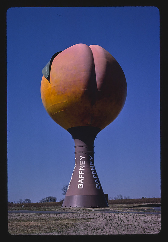 This vibrant image showcases the iconic Peachoid water tower in Gaffney, South Carolina, standing proudly against a brilliant blue sky. The colossal peach, with its distinct pink and orange hues and prominent crease, appears almost cheeky, a playful sentinel over the flat, open landscape. Its base boldly proclaims "GAFFNEY" in white lettering, anchoring this whimsical landmark. The background, a sparse field with distant trees, serves to highlight the Peachoid's unique and monumental presence, making it an unmistakable beacon for the town. This famous structure, also recognized from the TV show *House of Cards*, exudes a humorous charm, playfully winking at observers with its suggestive form.