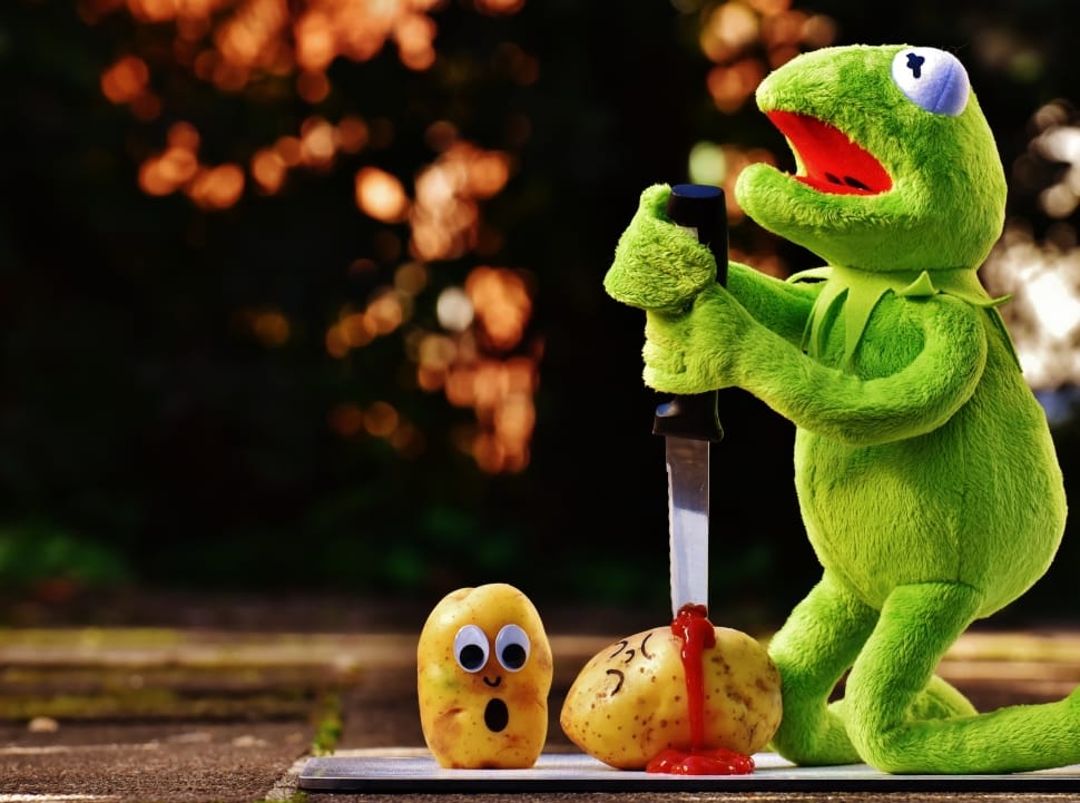 In this amusing scene, Kermit the Frog, the beloved Muppet character, is depicted in a hilariously surreal moment, wielding a knife over two animated potatoes. One potato looks shocked, with wide eyes and an open mouth, while the other appears to be "injured," oozing a comical red substance. Kermit's expression is one of determination, emphasizing the absurdity of the situation.

The background is softly blurred with warm bokeh lights, creating a contrasting cheerful atmosphere to the darkly humorous act taking place. This scene cleverly plays on internet meme culture, showcasing Kermit in an unexpected, absurd context, blending humor with a touch of surrealism. The juxtaposition of a friendly character like Kermit engaging in such an over-the-top action with vegetables adds to the comedic effect, making it both whimsical and entertaining.