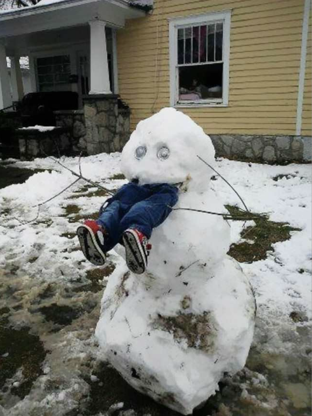 This humorous image features a two-tiered snowman, comically appearing to have "eaten" a child, whose legs in blue jeans and red-and-white sneakers dangle playfully from its midsection. The snowman's head sports two shiny, round objects, possibly car headlights, for eyes, giving it a wide-eyed, almost surprised demeanor, while thin branches serve as rudimentary arms. The ground is covered in patchy, melting snow, suggesting a recent snowfall or the tail end of winter. In the background stands a charming yellow house with white trim and a stone foundation, featuring a welcoming porch. The playful scene suggests a child's imaginative winter fun, perhaps a game of hide-and-seek gone wonderfully wrong, or a creative way to pose with their snowy creation, adding a lighthearted, whimsical touch to the snowy residential setting.