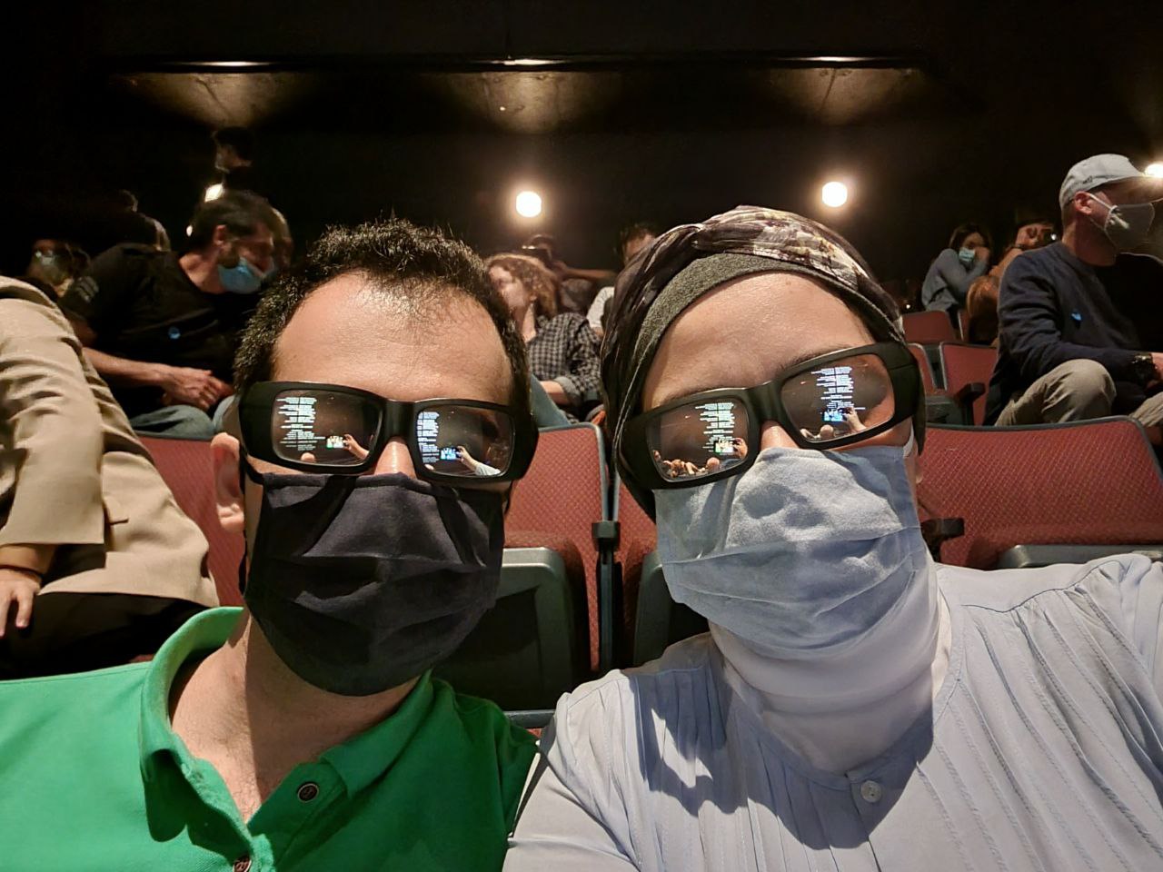 The image captures two individuals sitting in a theater, wearing 3D glasses and face masks, suggesting they are about to watch a movie. The man on the left has short, dark hair and is dressed in a green shirt, while the woman on the right wears a light blue top and a patterned headscarf. Their demeanor appears playful, as they both smile slightly, perhaps enjoying the anticipation of the film.
In the background, other audience members are also visible, many of whom are wearing masks, indicating a response to health guidelines. The dark setting of the theater adds a cozy and intimate atmosphere, perfect for movie-watching. The reflections in their glasses show snippets of the film or perhaps the opening credits, heightening the excitement.
This scene humorously captures the post-pandemic movie experience, blending safety with enjoyment, making it a relatable snapshot for many.