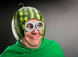 This humorous image features a person, likely male, with a mischievous smirk, looking slightly to the side. They are adorned in an absurd, homemade costume. A large watermelon, hollowed out and carved, serves as a helmet, complete with a small stem curling upwards at the back. Their eyes are covered by oversized, grey, cylindrical goggles, reminiscent of a Minion character, adding to the comical effect. The individual is draped in a vibrant green fabric, complementing the watermelon's color. The plain, dark grey background provides no specific context, allowing the viewer's full attention to be drawn to the subject's playful demeanor and the whimsical, fruit-inspired attire. The entire scene exudes a lighthearted and silly vibe.