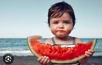 A charming baby with dark hair and striking blue eyes is captured mid-snack on a sunny beach. Dressed in a striped top, the little one holds a massive, vibrant red watermelon slice, almost as big as their torso. Their cheeks and chin are adorably smeared with red watermelon juice, a testament to their enthusiastic eating. Despite the messy evidence of a delicious treat, the baby maintains a surprisingly serious and contemplative expression, which adds a humorous contrast to the scene. The background features a serene blue ocean meeting a clear sky, with dark sand in the foreground, perfectly setting the stage for a quintessential summer day. The image humorously captures the pure, uninhibited joy of a child's summer indulgence.