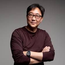 Actor Ke Huy Quan, celebrated for his roles in 'The Goonies' and 'Everything Everywhere All at Once,' beams with a confident, approachable smile, looking directly at the camera. His arms are crossed, conveying a relaxed yet assured demeanor, amplified by his stylish glasses. He sports a dark maroon long-sleeved shirt and a watch. The plain gray background serves as a minimalist stage, ensuring all focus remains on his engaging expression. It's a portrait of a seasoned performer, exuding a quiet charm and the wisdom of a remarkable career, perhaps contemplating his next cinematic triumph or simply enjoying the moment.