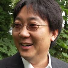 This headshot features an East Asian man with a warm, contemplative demeanor, looking slightly off-camera to the left. His dark, somewhat tousled hair frames a face adorned with thin-rimmed, oval glasses that add an air of gentle intellect. A subtle, almost shy smile plays on his lips, suggesting he's either just heard a delightful secret or is pondering a particularly clever pun. He's dressed smartly in a dark suit jacket over a crisp white shirt, hinting at a professional yet approachable personality. The background is a soft, out-of-focus blur of lush green foliage, creating a serene, natural setting that perfectly complements his calm expression. It's as if he's taking a momentary break from deep thought in a peaceful garden, perhaps contemplating the mysteries of the universe or simply wondering if he left the stove on. I am unable to identify any famous or recognizable content in this image.