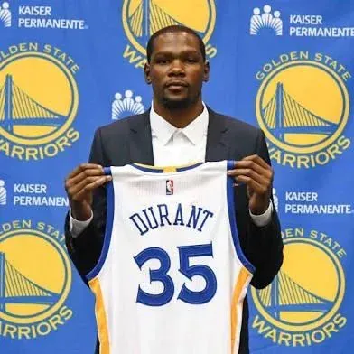 Kevin Durant, the renowned NBA player, is captured in a formal portrait, holding up a white Golden State Warriors jersey with 'DURANT' and the number '35' prominently displayed in blue. He wears a dark suit jacket over a crisp white shirt, presenting a serious and focused demeanor. The background is a vibrant blue banner featuring multiple Golden State Warriors logos, which depict the iconic Golden Gate Bridge in yellow, along with 'Kaiser Permanente' sponsorship branding. This image marks his official introduction and signing with the Golden State Warriors, a monumental event in NBA history that significantly shifted the league's power dynamics. The background directly contextualizes this pivotal moment, highlighting his transition to the team.