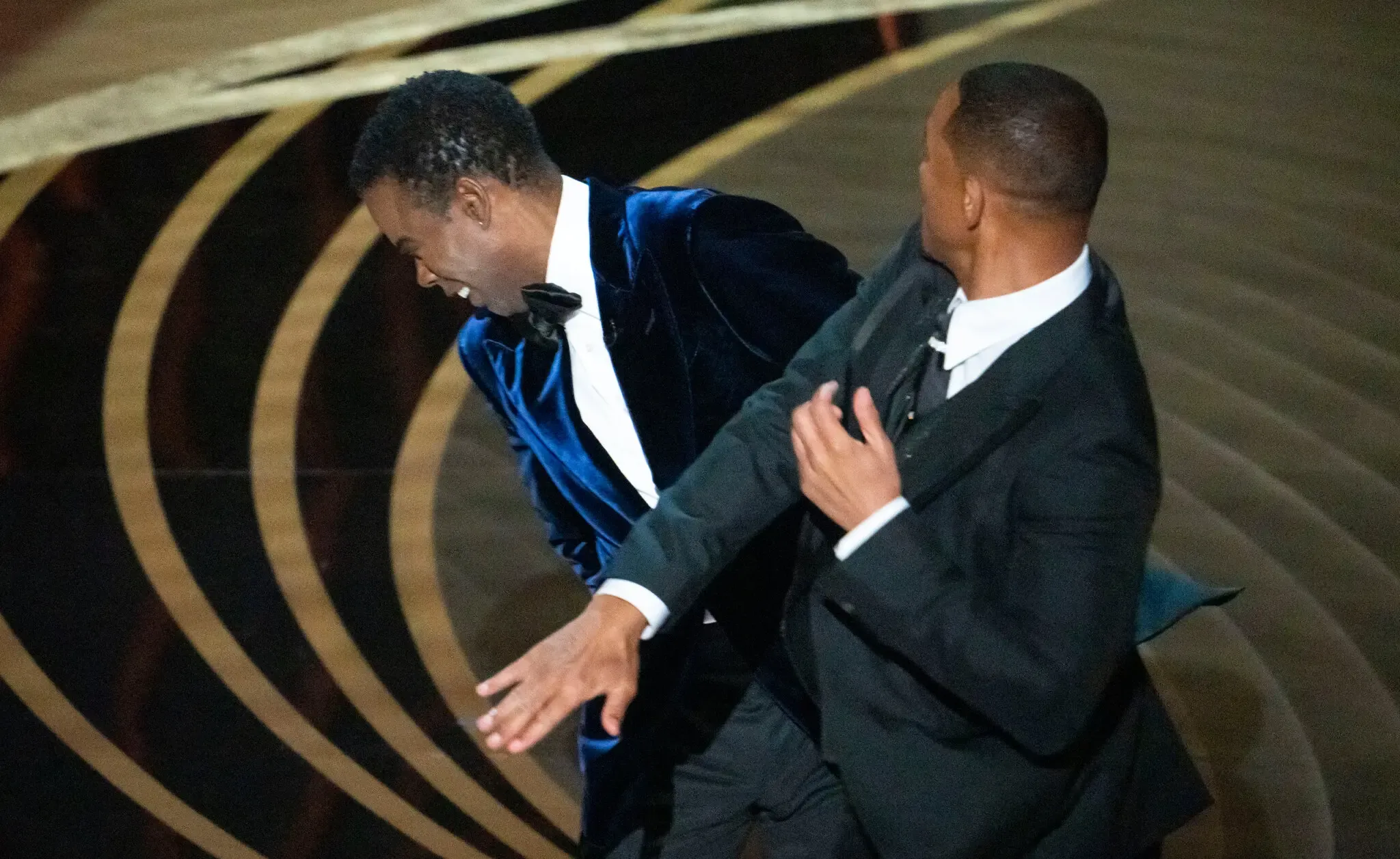 This image captures a pivotal moment from the 2022 Oscars, where Chris Rock, dressed in a sharp black tuxedo with a bow tie, is caught mid-laugh, seemingly surprised by Will Smith's unexpected action. Will Smith, in a classic black suit, is depicted just after delivering a slap, with his hand in motion, suggesting a mix of anger and determination. The background features the iconic Oscars stage, adorned with elegant golden swirls, which contrasts sharply with the tension of the moment. The audience's reaction is palpable, amplifying the image's significance in pop culture. This incident sparked widespread discussions about comedy boundaries and personal offense, making it a memorable Oscars moment. The absurdity of the situation, where humor turns to confrontation, adds a layer of dark comedy, highlighting the unpredictable nature of live events.