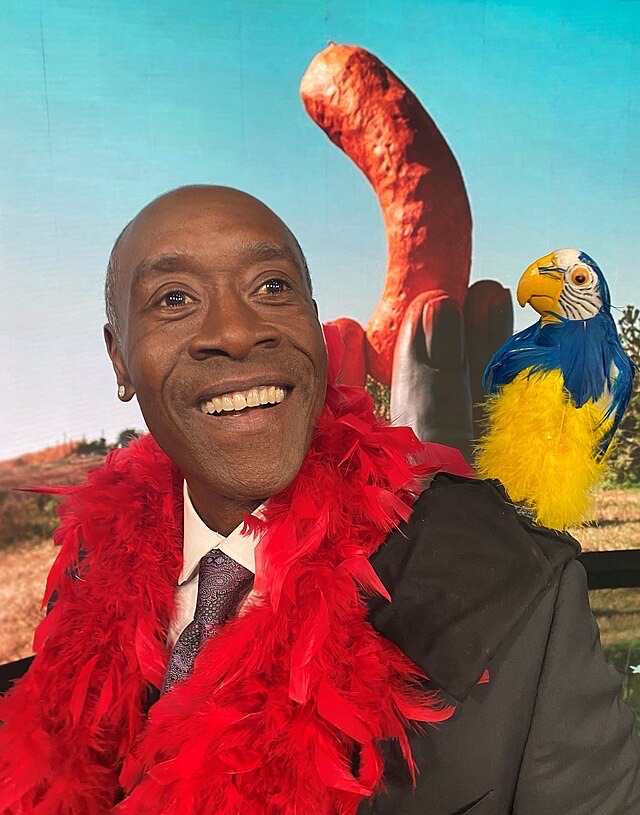 The image features Don Cheadle, radiating joy with a broad smile, adorned in a vibrant red feather boa that adds a playful flair to his appearance. Perched on his shoulder is a colorful parrot, its bright blue and yellow feathers contrasting beautifully with the background.
Behind them, a whimsical backdrop showcases a large, oddly-shaped rock formation that adds a humorous touch, suggesting a lighthearted or promotional event. The overall scene feels festive and carefree, evoking a sense of fun. Cheadle's demeanor is warm and engaging, as if he's enjoying a moment filled with laughter and camaraderie.
This delightful mix of vibrant colors, playful accessories, and Cheadle's infectious smile creates a memorable and humorous image, perfect for capturing the spirit of a lively event.