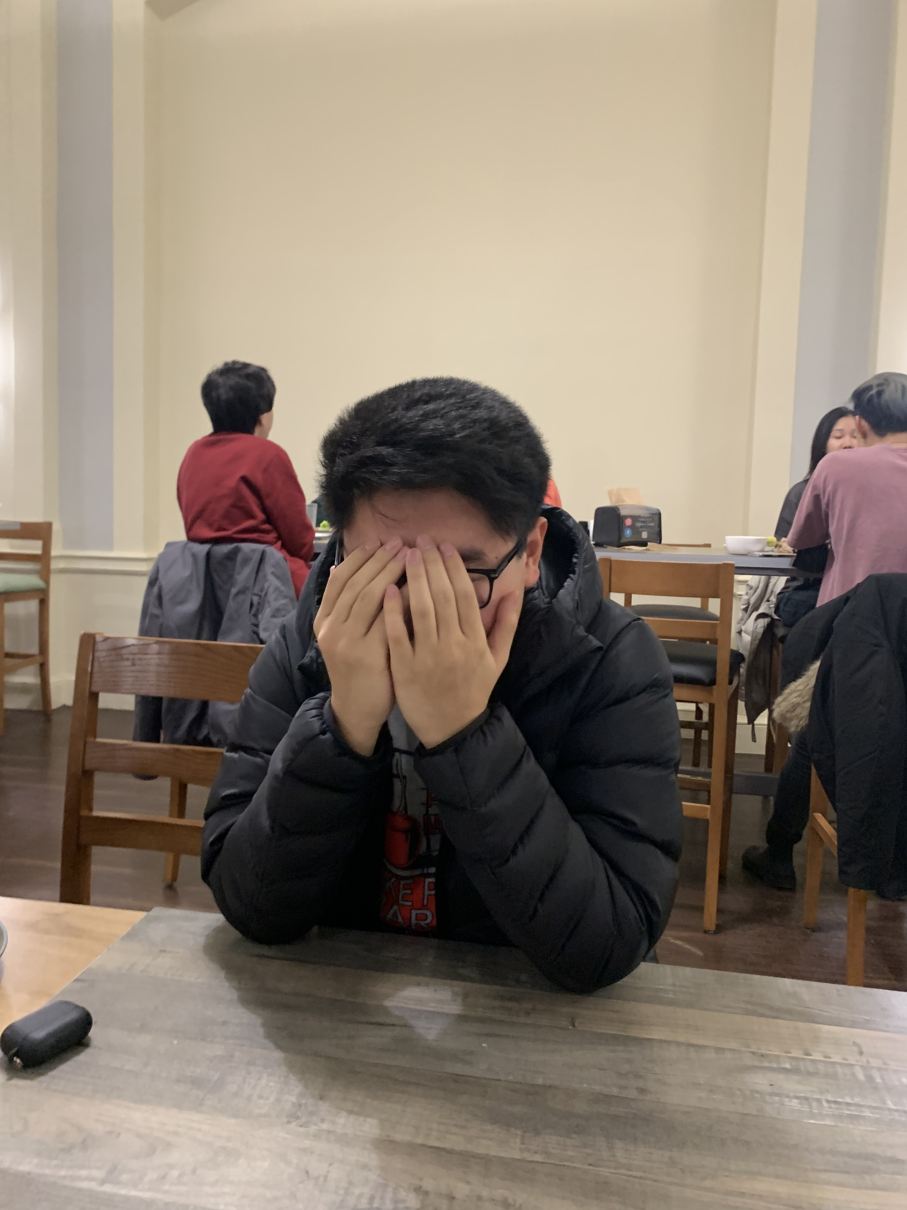 In the image, a young person is seated at a wooden table, their demeanor suggesting a mix of frustration or embarrassment, as they cover their face with both hands. They wear a dark puffer jacket, hinting at a casual and possibly chilly environment. The background features other diners, indicating a social setting, likely a restaurant or café, creating a relatable ambiance of shared experiences.

The lighting is warm, contributing to a cozy atmosphere, while the wooden furniture adds a rustic charm. The subject’s pose could evoke humor, reminiscent of those moments when one just wants to disappear, perhaps due to an awkward situation or a joke gone wrong. 

The scene captures the essence of youthful vulnerability, making it both relatable and amusing, as many can identify with the urge to hide in moments of embarrassment.