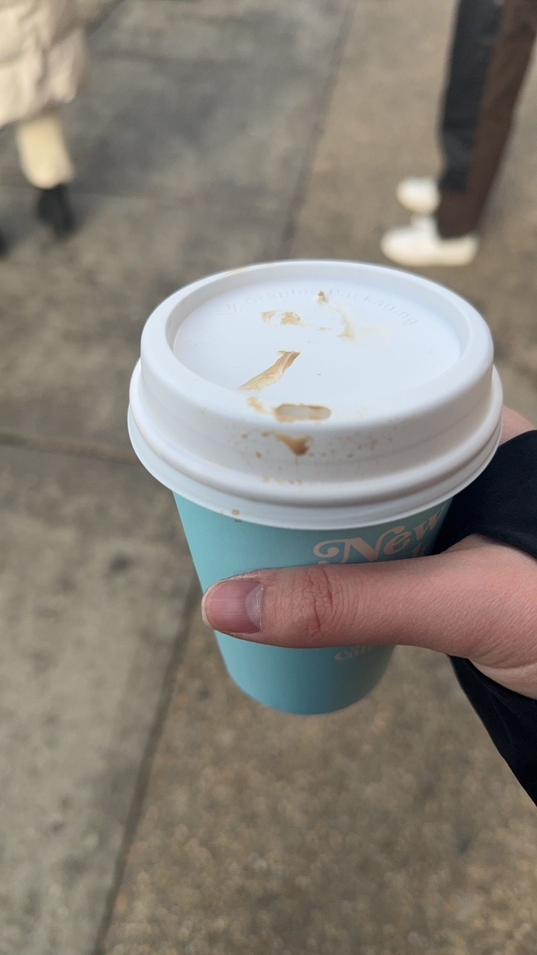 This close-up image features a hand holding a light blue coffee cup with a white lid, adorned with abstract, dried coffee stains that humorously resemble a tiny, caffeinated Rorschach test. The cup, possibly from 'New' (partially visible text), suggests a casual, on-the-go moment. The blurry background reveals a paved sidewalk and the legs of other pedestrians, reinforcing the bustling urban context. The slightly messy lid hints at the everyday reality of a quick coffee run, perhaps a minor skirmish with a stubborn lid or a hurried sip. It's a relatable snapshot of daily life, where even a simple coffee cup tells a story of movement and morning rituals.
