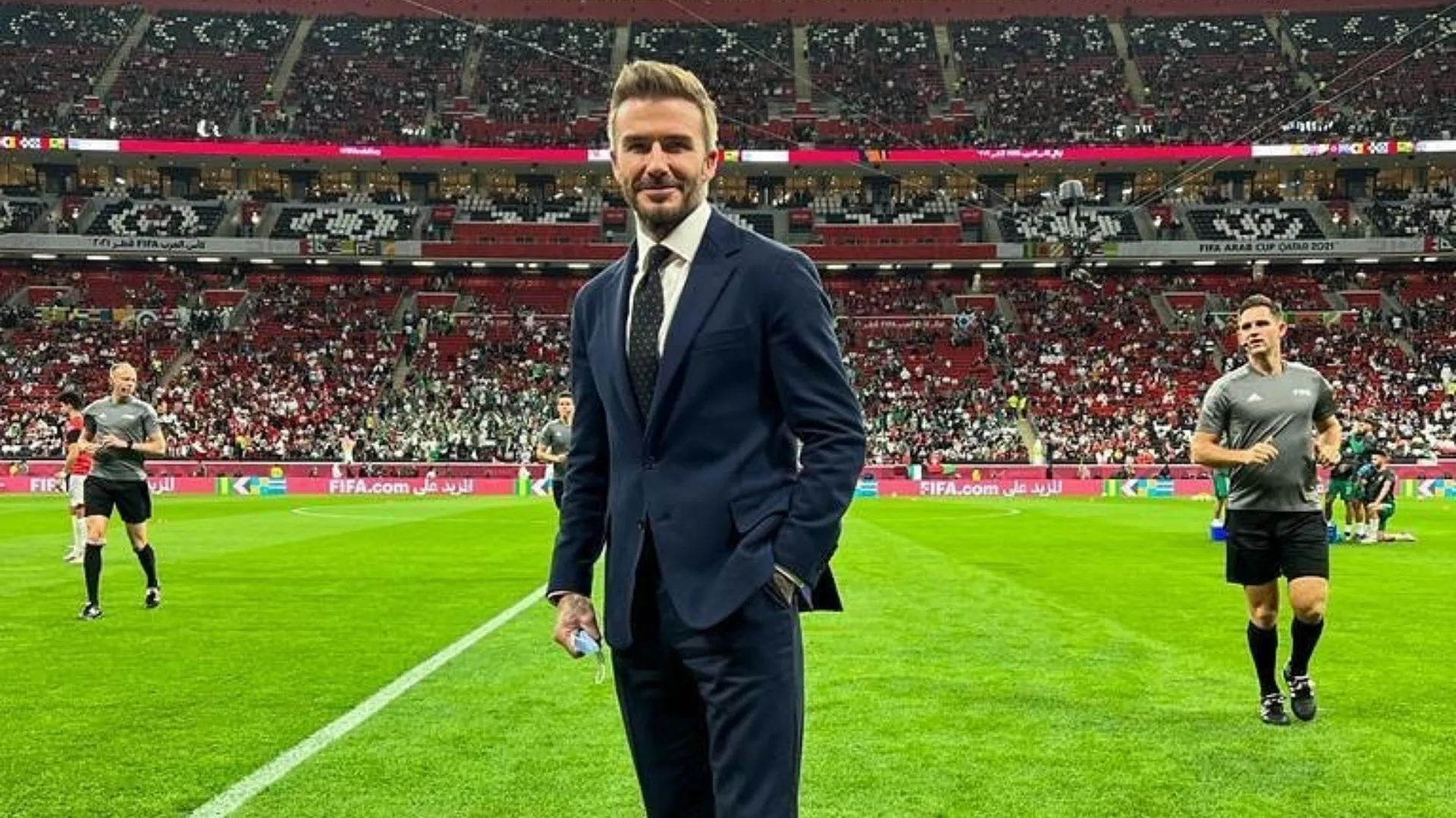 The image captures David Beckham at a FIFA World Cup event, exuding confidence and charm in a sleek navy suit. His demeanor is relaxed yet composed, a slight smile hinting at his enjoyment of the moment. The lush green pitch contrasts with the vibrant red and white of the enthusiastic crowd in the background, creating an energetic atmosphere.

The stadium, likely a prominent venue in Qatar, buzzes with excitement, filled with fans ready for the match. Beckham stands in the foreground, embodying the spirit of football and its global appeal. He seems to be a bridge between the past and present of the sport, having transitioned from a player to an ambassador.

Humorously, one might think Beckham, in his sharp suit, could be mistaken for a footballing CEO, ready to negotiate a transfer deal right from the pitch!
