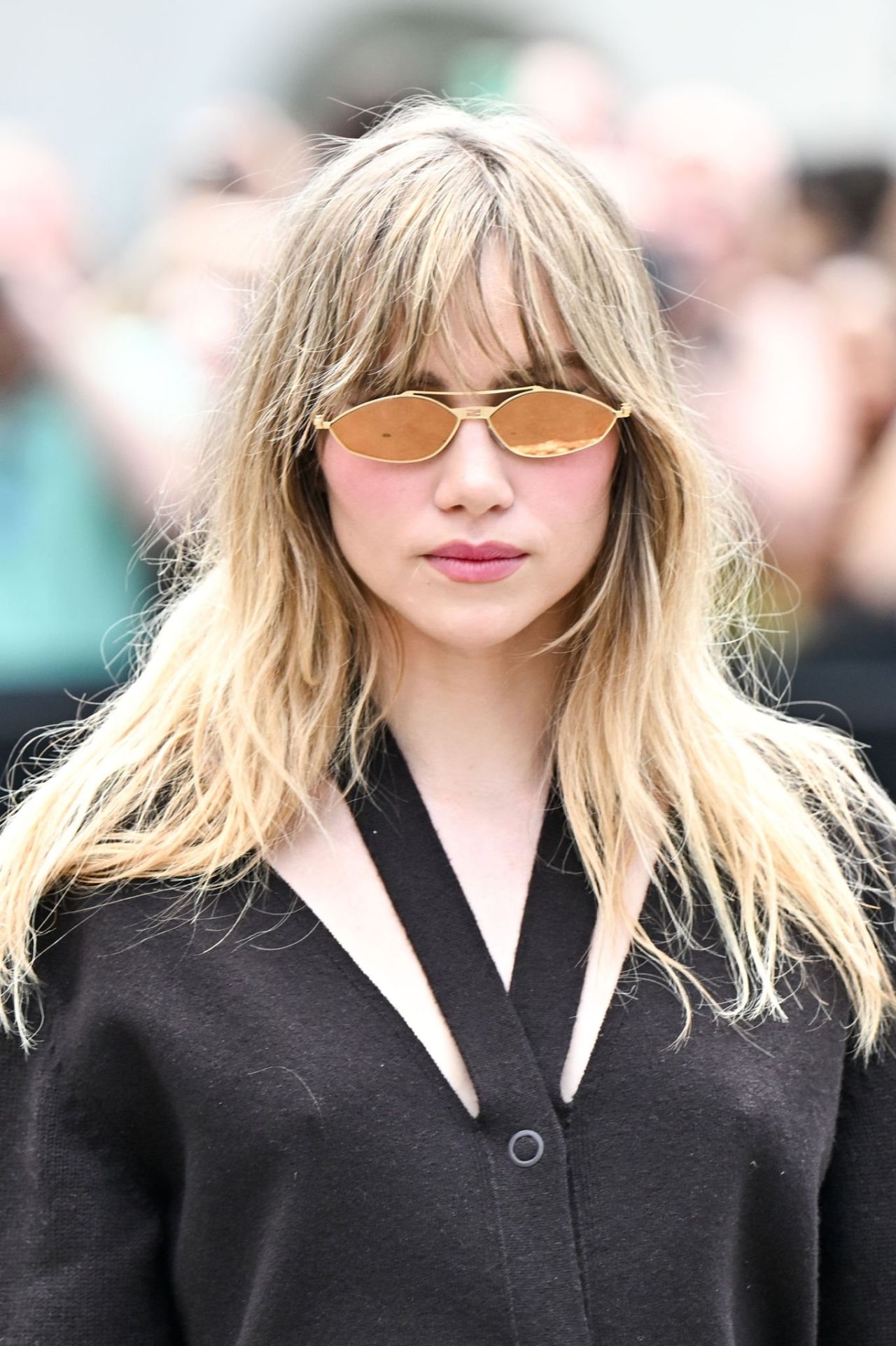 The image features a stylish individual with long, tousled hair, wearing unique gold-tinted sunglasses that lend a chic and confident air. Her expression is serene yet assertive, suggesting she is comfortable in her surroundings, perhaps even enjoying the attention.
The subject is dressed in a dark, elegant outfit with a distinctive neckline, adding to her fashionable presence. The background appears to be a crowd, with blurred figures hinting at a busy event, possibly a fashion show or a public appearance, where her style is on full display.
This context enhances the image, making it feel lively and dynamic. The overall vibe is one of effortless cool, as if she's a trendsetter casually navigating the spotlight. One might imagine her thinking, “These sunglasses? Oh, they’re just for keeping the paparazzi at bay.”