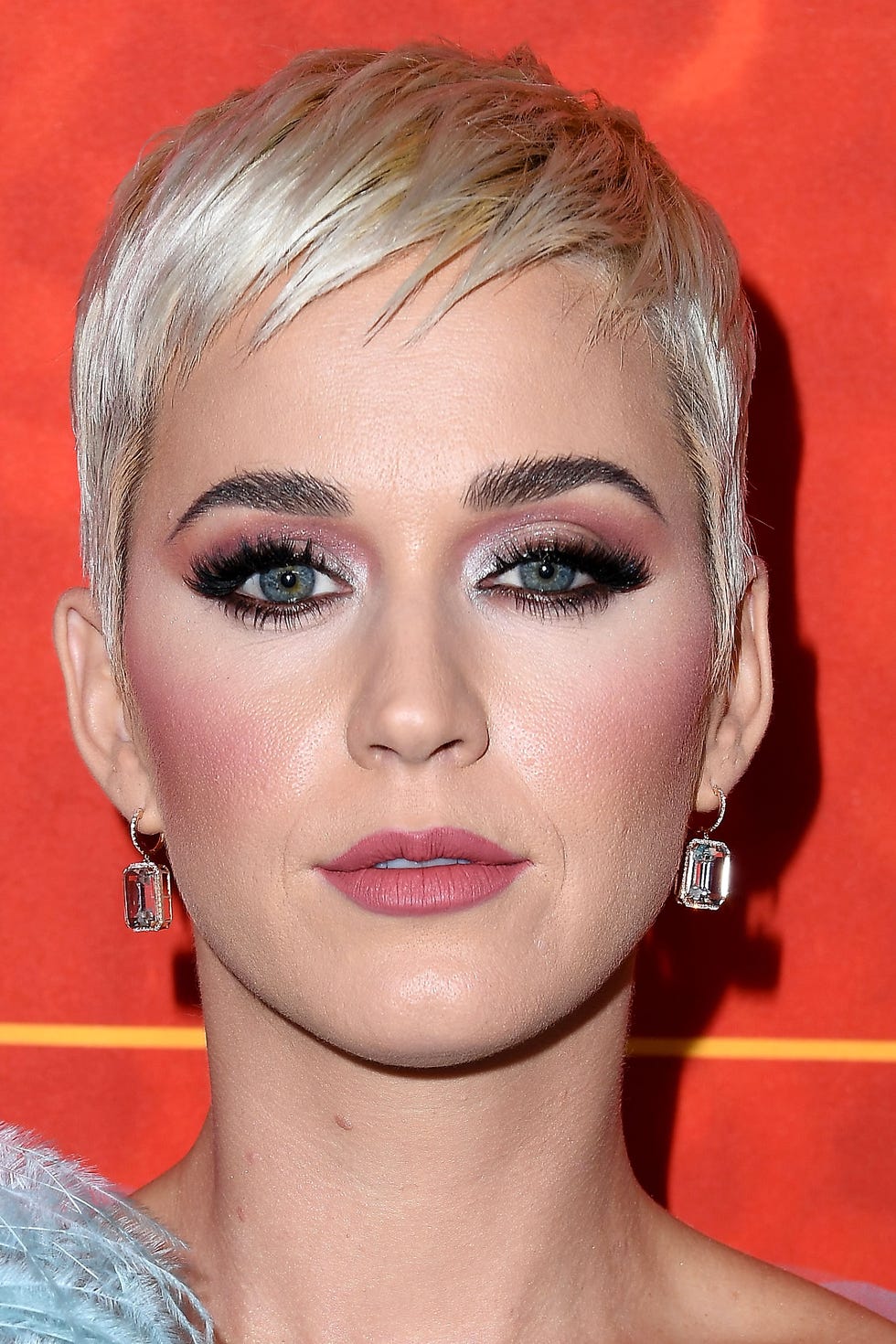 The image features a close-up of Katy Perry, showcasing her striking makeup and signature short blonde hair. Her demeanor appears confident and poised, with a subtle expression that suggests she's in the spotlight, possibly at a glamorous event. The bold eye makeup, complemented by long lashes, draws attention to her expressive eyes, while her soft pink lips add a touch of elegance.

The background is a vibrant red with a hint of orange, creating a warm and inviting atmosphere that enhances her radiant look. This color choice likely signifies excitement and energy, fitting for a public appearance by a pop icon known for her lively performances.

Overall, this image captures Katy Perry's iconic style and persona, making it a vivid representation of celebrity glamour. It's as if she’s ready to burst into song, or perhaps drop a witty remark about the latest fashion trends!