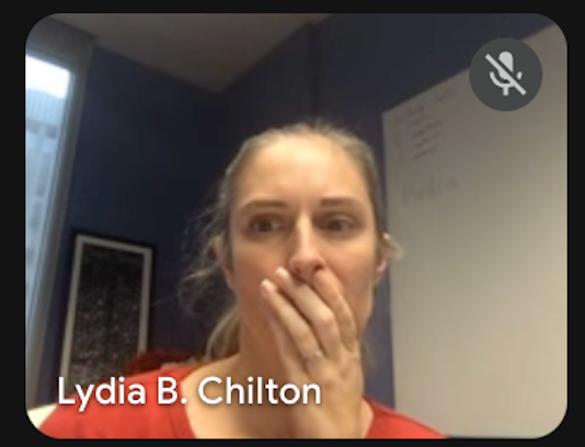 The image captures Lydia B. Chilton during a video call, her expression a dramatic mix of wide-eyed surprise and perhaps a stifled gasp, with her hand covering her mouth. She appears to have just witnessed or heard something utterly astonishing. The muted microphone icon in the top right corner adds a humorous touch, suggesting her reaction is silent but profound, perhaps saving her colleagues from an audible outburst. The background is a simple office setting with a dark blue wall, a framed picture, and a whiteboard with faint writing, typical of a remote work environment. Her intense reaction contrasts humorously with the mundane setting, making one wonder what incredible revelation just unfolded on screen.