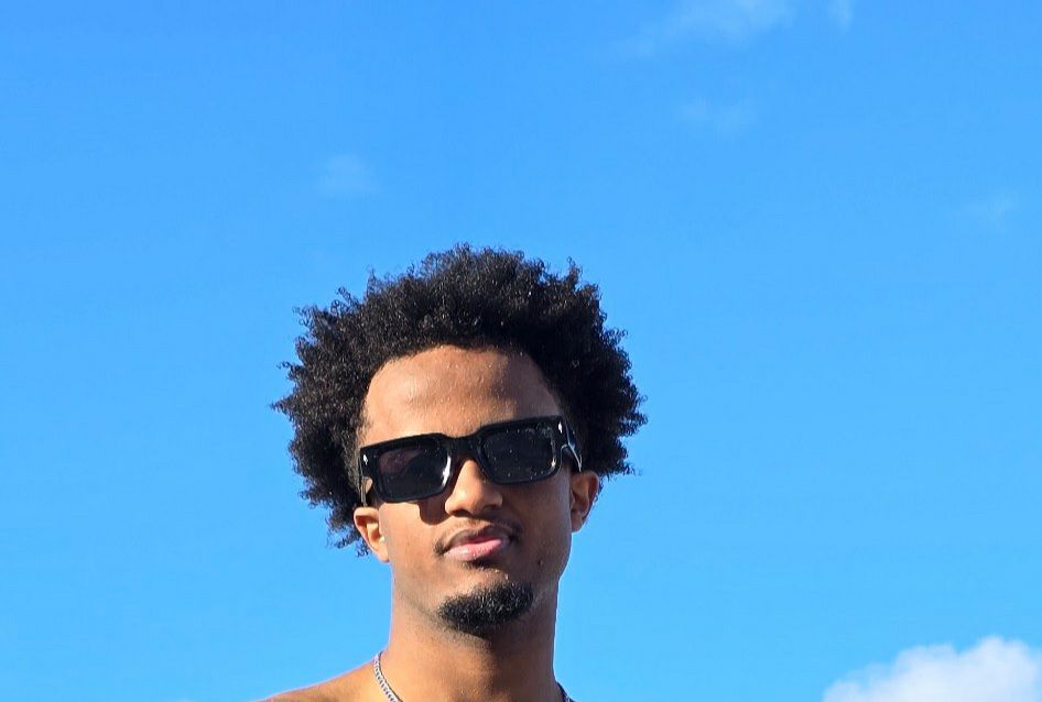 The image features a young man with a confident demeanor, sporting stylish sunglasses and a relaxed expression. His curly hair frames his face, adding to his cool vibe. The bright blue sky in the background suggests a sunny day, enhancing the carefree, summer-like feel of the scene.

The absence of any prominent buildings or specific landmarks hints that this could be a casual outdoor setting, perhaps at the beach or a park, where one might enjoy leisure activities. The overall mood is light-hearted and fun, perfect for capturing a moment of enjoyment.

Humorously, one might imagine the sunglasses as a shield against the sun's rays, making him feel like a celebrity on a secret mission to find the best ice cream in town. The carefree atmosphere invites smiles, making it a snapshot of a perfect day.