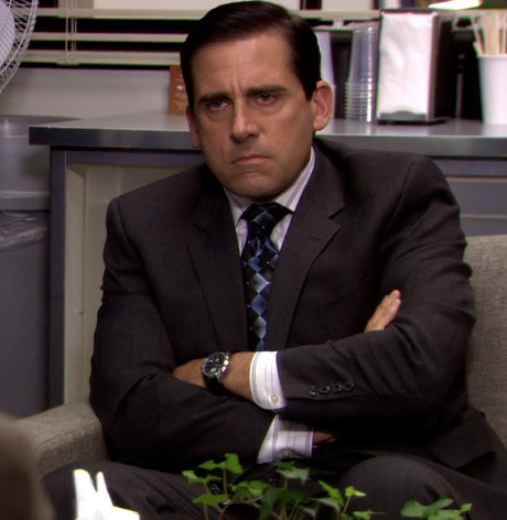 The image features Steve Carell as Michael Scott, seated at his desk in the office of Dunder Mifflin, the fictional paper company from the TV show 'The Office'. His demeanor is serious and contemplative, a stark contrast to his usual bumbling and humorous personality. The expression on his face suggests he is deep in thought or perhaps frustrated with a situation at work.
In the background, we see typical office elements like a fan and neatly arranged office supplies, reinforcing the mundane yet chaotic atmosphere of corporate life. This setting is integral to the show, as it highlights the often absurd scenarios that unfold in a typical day at the office. The image captures a moment of unexpected seriousness, humorously juxtaposed with the lighthearted tone of the series.