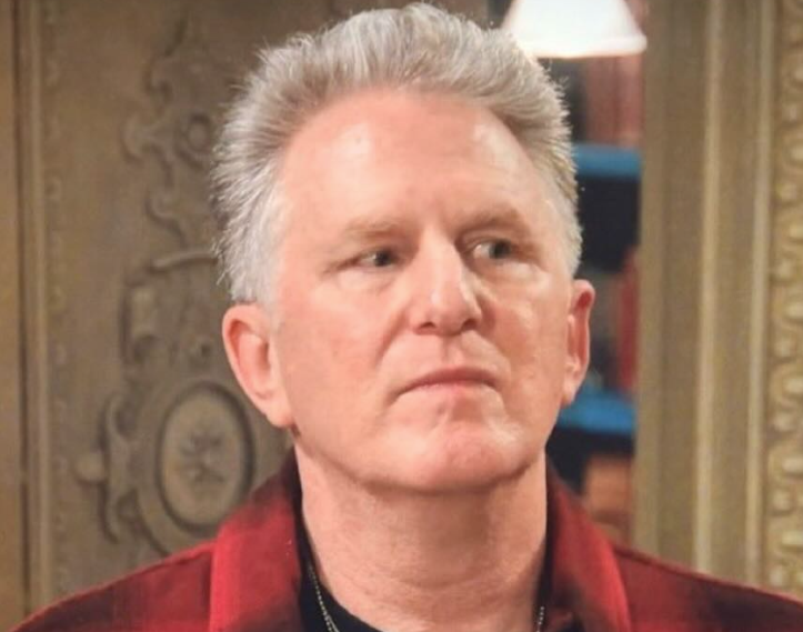 This close-up image features actor Stephen Collins, known for his role as Reverend Eric Camden in '7th Heaven,' sporting neatly styled grey hair and a red and black plaid shirt over a dark t-shirt. His demeanor is one of focused contemplation or mild skepticism, as he gazes intently upwards and to the right with a neutral, closed-mouth expression. The background, though slightly blurred, reveals an ornate, possibly carved wooden panel on the left, suggesting a classic interior. On the right, hints of bookshelves and a light fixture are visible, indicating a well-appointed indoor setting, likely a set from a television show or movie. The background elements complement the foreground, creating a sense of a traditional, perhaps cozy, yet slightly formal environment. His intense side-glance humorously suggests he's either just heard a shocking secret or is silently judging your life choices.