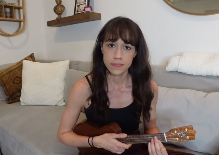 Colleen Ballinger, known for her internet character Miranda Sings, is captured in a medium shot, seated on a light grey couch while holding a ukulele. She wears a black tank top and has long, dark hair with bangs. Her expression is notably serious and somewhat deadpan, which, given her comedic background, adds a subtle layer of humor, perhaps hinting at her Miranda Sings persona's earnest (yet comically bad) musical endeavors. The background reveals a cozy home setting, typical of a YouTuber's content, featuring decorative pillows, a floating shelf with a bust and framed photos, and a round mirror. This familiar domestic backdrop reinforces the casual, direct-to-camera style often seen in her videos, where the ukulele is a frequent prop.