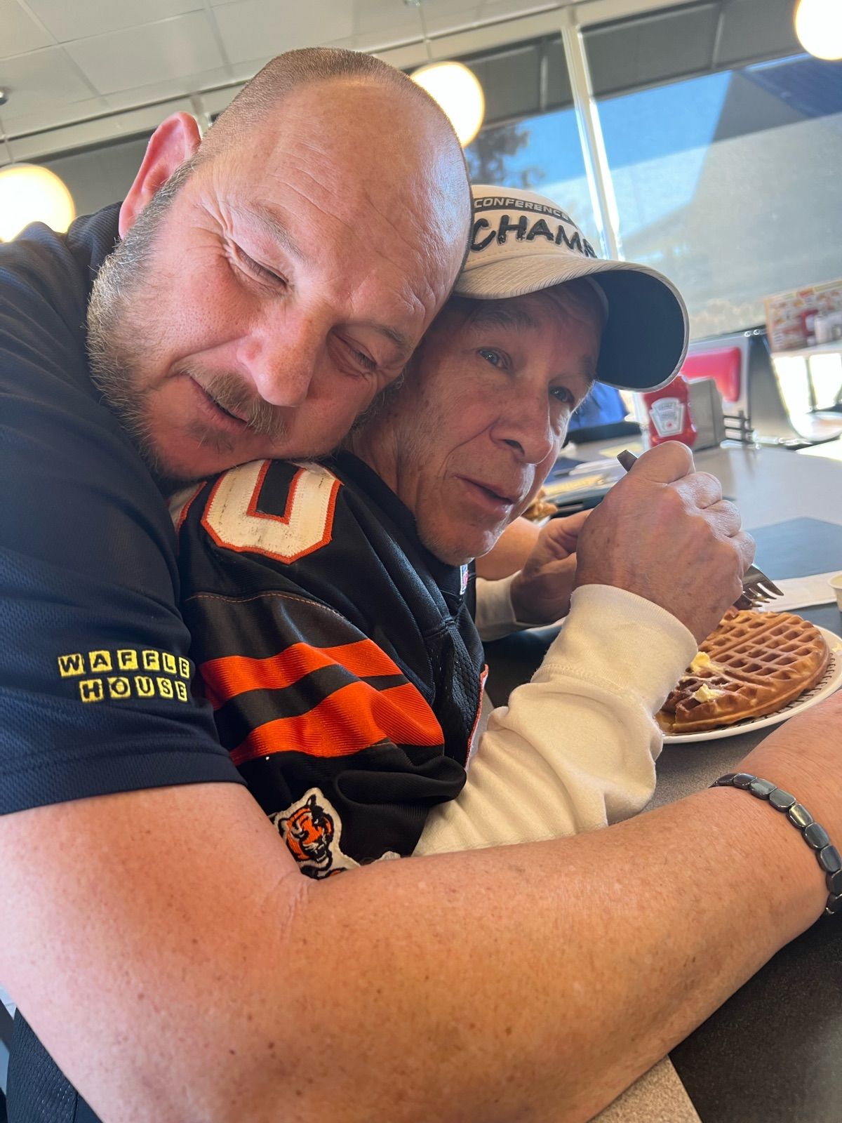 Two men share an affectionate moment inside a Waffle House. The larger man, wearing a black polo with 'WAFFLE HOUSE' on the sleeve, embraces the other man from behind, eyes closed in what appears to be pure contentment or perhaps a post-celebration exhaustion. The man being hugged, sporting a 'CONFERENCE CHAMP' hat and a black and orange striped jersey with a tiger logo, looks directly at the camera with a slightly surprised but accepting expression, fork poised over his waffle. The background clearly shows the interior of a Waffle House, with windows and diner-style seating, perfectly complementing the man's shirt and the waffle on the table. The humor lies in the intense, almost smothering hug contrasting with the casual act of eating a waffle, suggesting a celebratory meal after a big win, perhaps for the 'Conference Champs'.