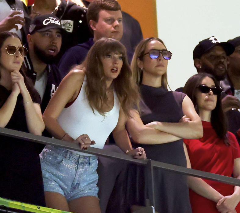 The image features Taylor Swift, known for her engaging stage presence and emotive performances, visibly expressing excitement or concern as she watches an event. Her demeanor, marked by wide eyes and an animated posture, indicates a strong emotional investment in what’s happening. She is dressed casually in a white top and sparkling denim shorts, embodying a relaxed yet stylish vibe.
Surrounding her are friends and fans, each sporting various expressions that suggest a mix of anticipation and enthusiasm. The background hints at a lively atmosphere typical of a concert or sports game, amplifying the overall excitement of the scene. The diverse crowd reflects the energy of a live event, where emotions run high. Taylor's presence, combined with the charged ambiance, captures a moment of connection with her fans and the thrill of live entertainment.