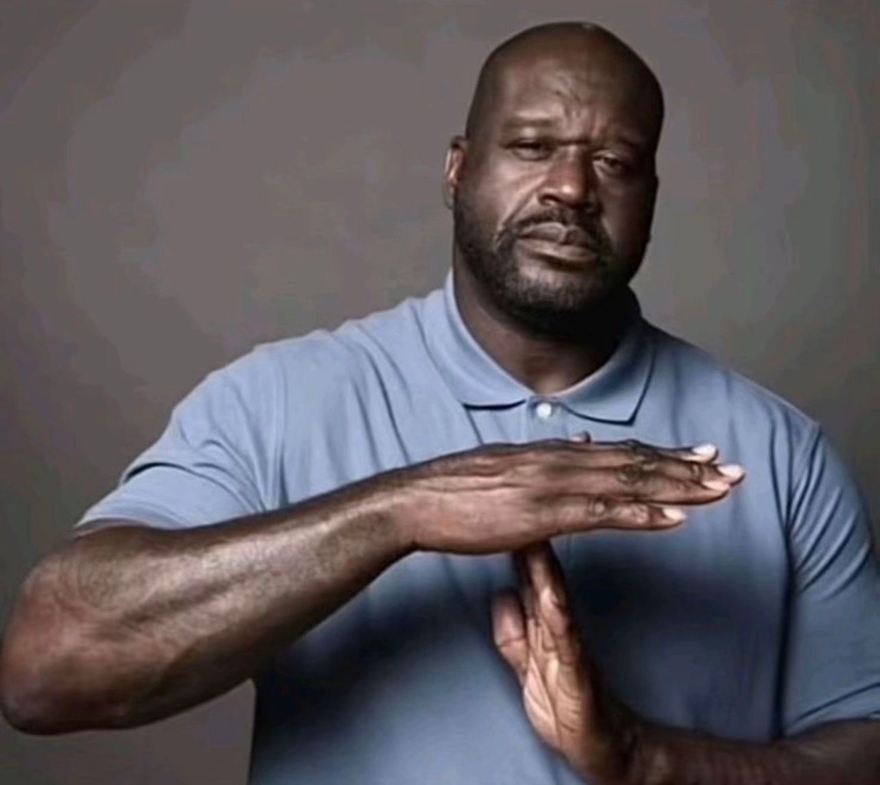 The image features basketball legend Shaquille O'Neal, known as Shaq, against a plain grey background. He wears a light blue polo shirt and displays a serious, almost exasperated expression, with furrowed brows and a slightly downturned mouth. His demeanor suggests a moment of frustration or a need for a pause. Shaq is prominently making the universally recognized "timeout" gesture with his hands, forming a 'T' shape. This iconic pose has become a popular meme, humorously used to signal a need to stop, take a break, or intervene in a chaotic situation. The simple background ensures all attention is on Shaq and his emphatic gesture, amplifying the comedic potential of his serious expression combined with the relatable "timeout" signal.