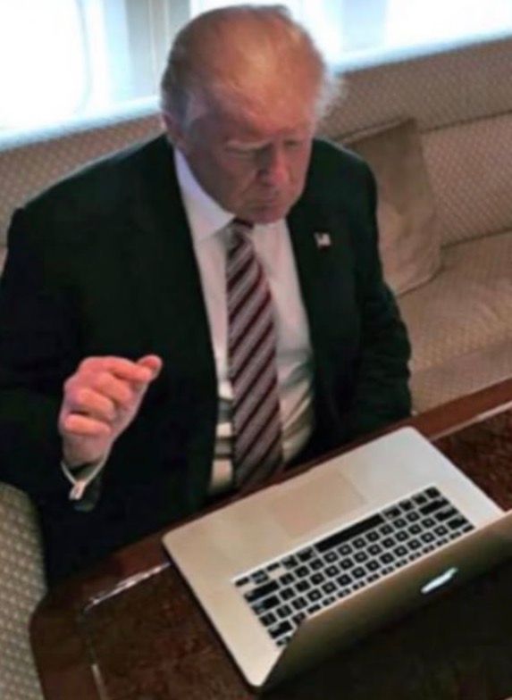 The image shows an older man with light-colored hair, wearing a black suit jacket, a white dress shirt, and a striped tie. He is seated at a table with a silver laptop in front of him. The man appears to be looking at the laptop screen with a focused expression. The background includes a cushioned bench or sofa with a pillow. The setting appears to be indoors, possibly in a formal or office-like environment.