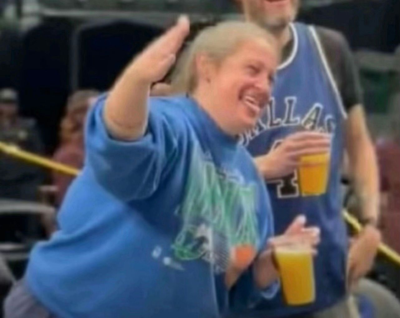 The image captures a lively moment featuring a woman in a bright blue sweatshirt, joyfully gesturing with one hand raised as if to emphasize a point or celebrate a moment. Her smile radiates happiness, suggesting she is fully engaged in the fun atmosphere around her. She holds a cup in her other hand, possibly containing a drink, which adds to the festive vibe.
In the background, there are blurred figures, hinting at a gathering, possibly at a concert or festival. The colors and casual attire suggest a relaxed, social setting. The man's presence behind her, also holding a drink, reinforces the social aspect, indicating camaraderie and enjoyment.
Overall, the scene exudes a sense of joy and carefree spirit, making it easy to imagine this as a snapshot of a fun event filled with laughter and celebration.