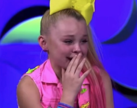 In this emotional moment, JoJo Siwa, a well-known dancer and singer, is captured with tears in her eyes, conveying a mix of vulnerability and resilience. She's wearing a bright pink outfit decorated with colorful accessories, highlighting her signature vibrant style. Her large yellow bow adds a touch of trademark flair, emphasizing her youthful energy. In the background, abstract shapes in soft colors create a supportive, theatrical atmosphere, often seen in reality TV settings, particularly on shows like 'Abby’s Ultimate Dance Competition.' This backdrop complements her emotional state, suggesting a dramatic moment of personal reflection or challenge. JoJo's demeanor strikes a balance between strength and sensitivity, making the image resonate with viewers who appreciate her journey and tenacity in the spotlight. Her expressive face captures the essence of a relatable struggle, embodying the trials of young performers.
