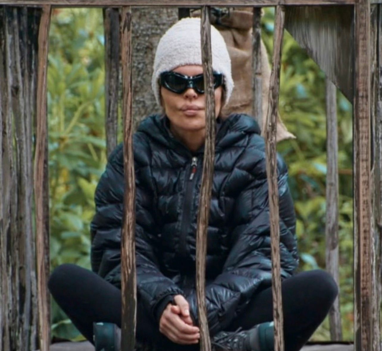 In the image, a person sits calmly behind wooden bars, dressed in a cozy black puffer jacket and a white knit hat. They wear dark sunglasses, adding an air of mystery to their demeanor. The subject's expression is neutral, perhaps reflecting a sense of contemplation or tranquility, as they sit cross-legged, seemingly at ease in their surroundings.
The background features lush greenery, emphasizing a natural setting that contrasts with the wooden structure in front of them. This setup could suggest a rustic cabin or a playful treehouse, evoking a whimsical atmosphere. The juxtaposition of the bars hints at confinement, yet the subject appears unbothered, almost embracing the moment.
This image might evoke humor, as the serious demeanor combined with the playful setting creates a funny contrast, as if they're playing a game of hide-and-seek but forgot they're not supposed to be behind bars!