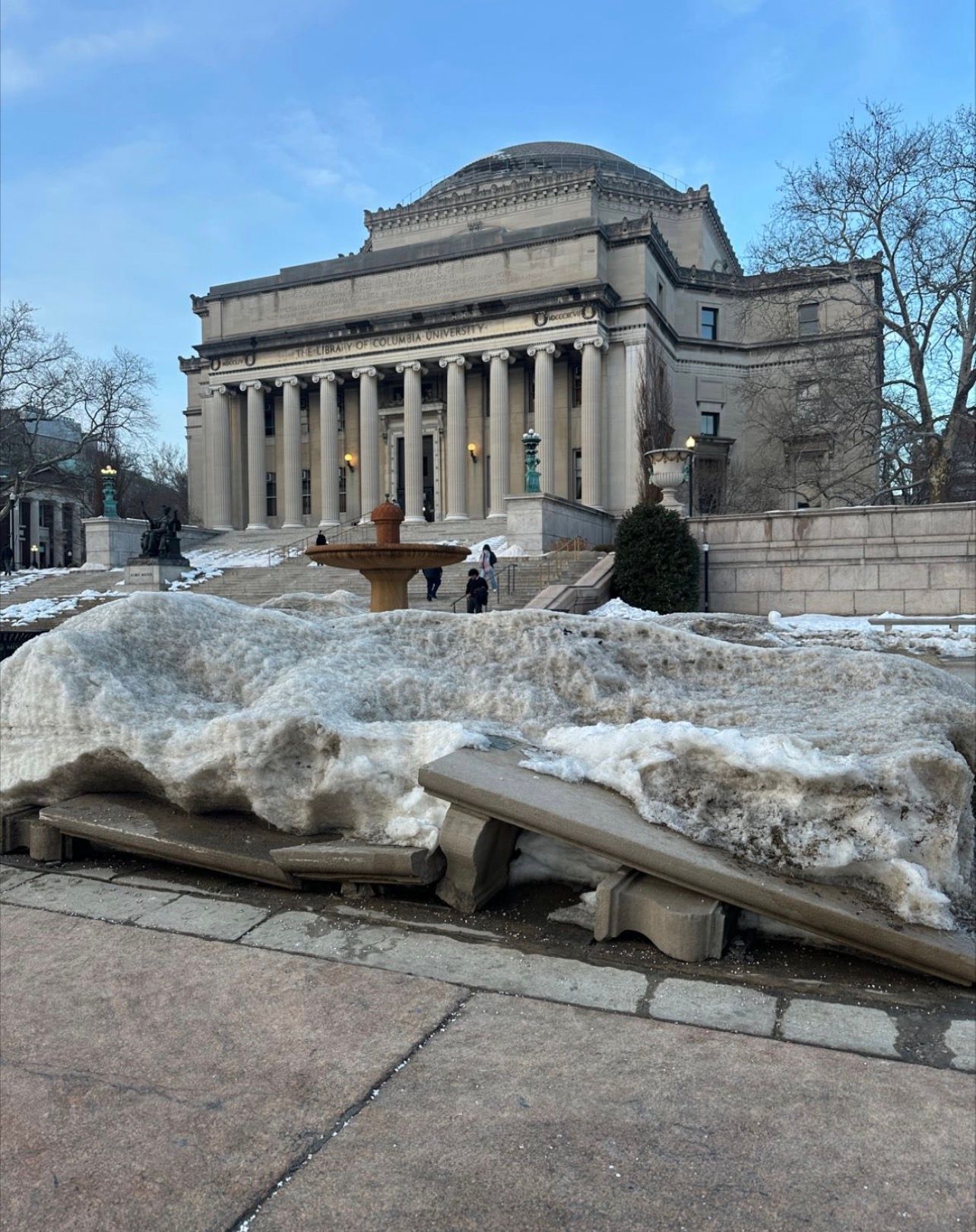 The image depicts the Library of Columbia University, an impressive architectural structure characterized by its grand columns and dome, conveying a sense of knowledge and academia. In the foreground, patches of snow and ice are melting, with a fountain partially obscured by the remnants of winter's chill. The demeanor of the scene feels contemplative, as a couple of people walk up the steps, perhaps in pursuit of inspiration or information.
The background serves as a stark contrast to the foreground's remnants of winter, highlighting the transition from cold to the promise of spring. The grand library stands as a beacon of learning amid the thawing landscape, suggesting renewal and growth. The juxtaposition of the enduring institution against the fleeting winter elements adds a layer of humor, reminiscent of nature's slow negotiation with time—like snow reluctantly yielding to the sun’s warmth.