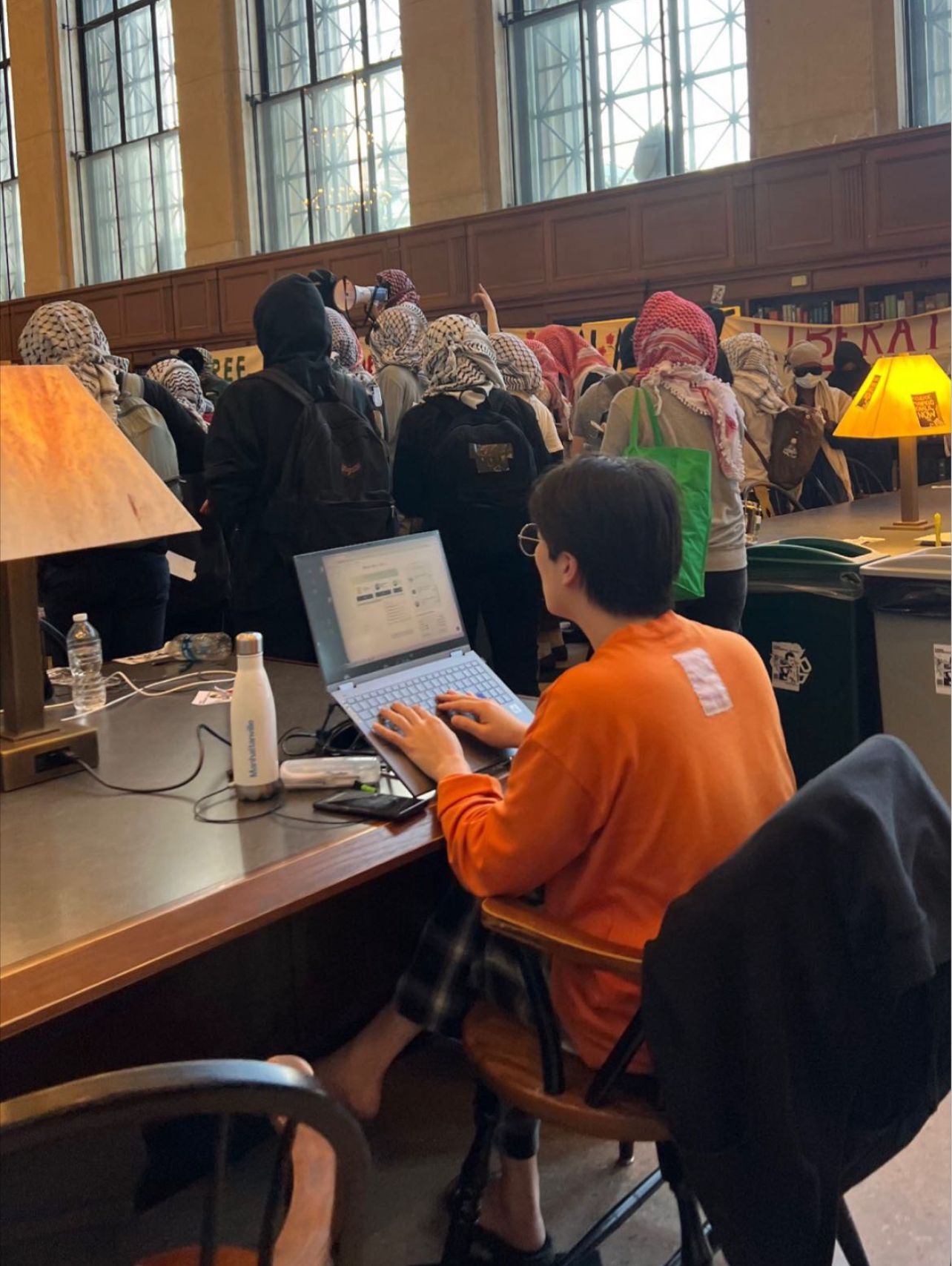 In a grand library hall, a person with short dark hair, wearing an orange shirt and plaid pants, is intensely focused on their laptop at a wooden desk. Their bare feet are visible, suggesting a relaxed demeanor. Behind them, a bustling crowd of people, many adorned with keffiyeh-style head coverings, are gathered, some holding a megaphone and banners. The background appears to be a lively protest or demonstration, contrasting sharply with the foreground subject's serene concentration. The humor lies in the individual's complete absorption in their work, seemingly unfazed by the energetic public gathering unfolding just feet away. This creates a comical juxtaposition of studious calm amidst passionate activism within an ornate academic setting.
