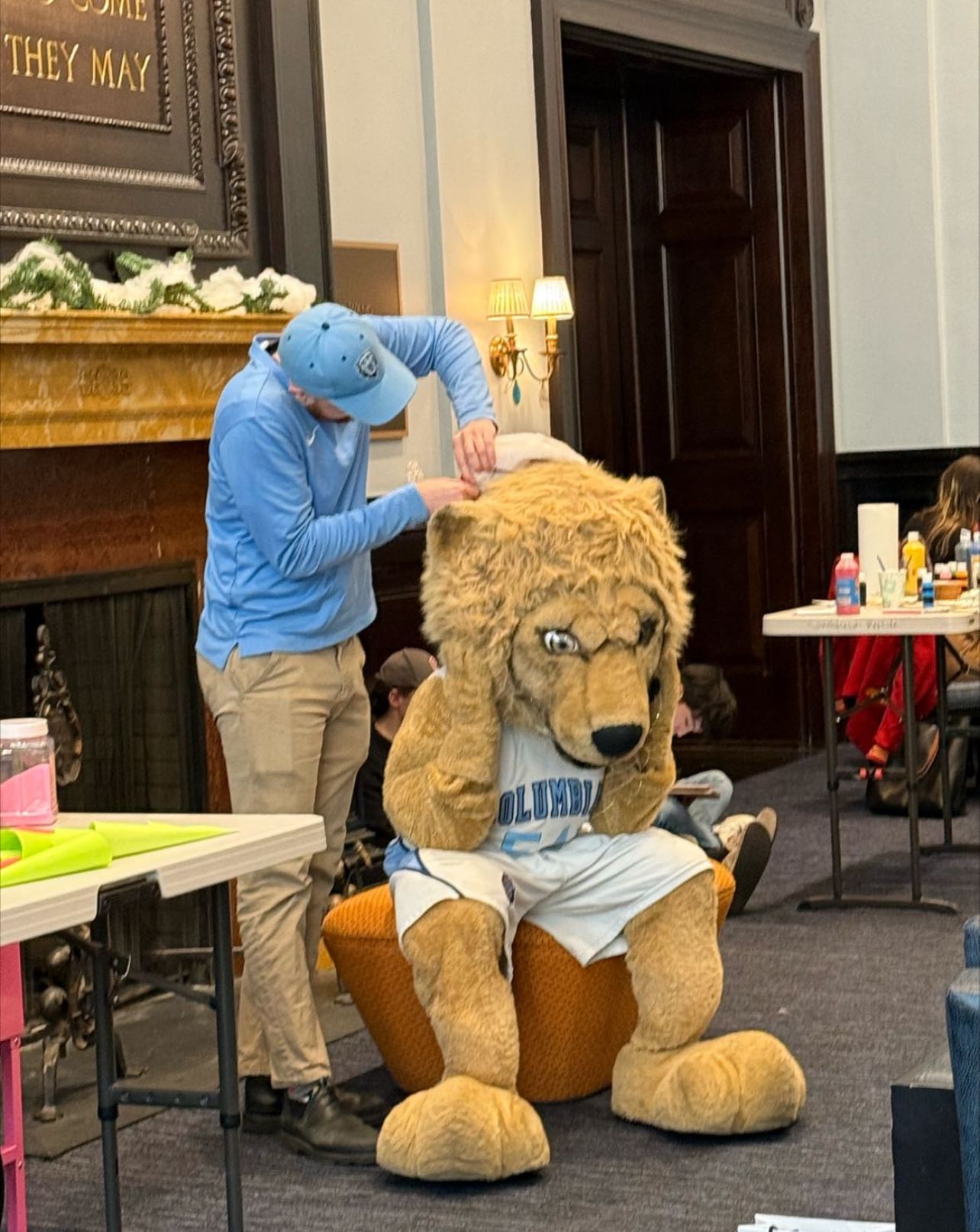 The Columbia University mascot, a lion in a "COLUMBIA" jersey and white shorts, sits on an orange pouf, comically holding its head as if suffering from a massive headache or a bad hair day. Its narrowed eyes suggest a demeanor of mild exasperation or discomfort. A handler, wearing a light blue hoodie and a matching Columbia cap, meticulously adjusts something on the mascot's head, perhaps an ear or part of the mane. The scene unfolds in what appears to be an elegant university hall, featuring a grand fireplace adorned with festive "snow" and garland, and a dark wooden door. The background also shows tables with craft supplies and other people, indicating a lively campus event. The humor lies in the fierce lion mascot looking utterly put-upon while being prepped, like a celebrity enduring a tedious styling session.
