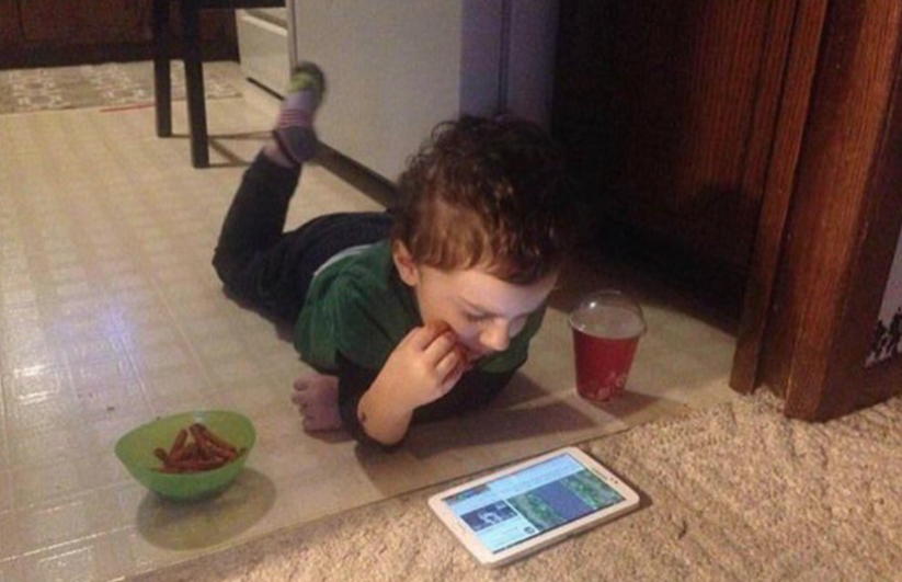 A young child with curly brown hair is completely engrossed in a white tablet, lying on his stomach on a light-colored tiled floor that transitions into a carpet. He's propped on his elbows, intently focused on the screen, with one bare foot and one foot in a striped sock playfully bent upwards. A green bowl filled with reddish snacks and a red plastic cup with a drink are conveniently placed beside him, indicating a full-on entertainment session. His demeanor is one of deep concentration, utterly lost in the digital world. The domestic background of a home setting perfectly complements this relatable scene of modern childhood indulgence. The humor lies in his complete dedication to the screen and snacks, a universal tableau of youthful escapism. I did not find any famous or recognizable content in this image.