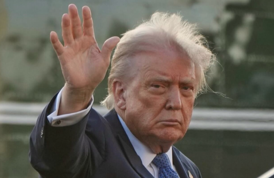 The image captures Donald Trump in a close-up, raising his right hand in a wave. Despite the friendly gesture, his demeanor appears serious, with a slightly furrowed brow and a stern expression. He is dressed in a dark suit, white shirt, and a blue patterned tie. His signature blonde-white hair is notably disheveled and windswept, particularly on top, adding an almost comical, windswept charm. The background is a blurred, indistinct mix of green and grey, strongly suggesting an outdoor setting, likely near an official vehicle such as Air Force One, a common backdrop for his public appearances. The humor arises from the contrast between his stern face and the waving hand, amplified by his famously unruly hair.