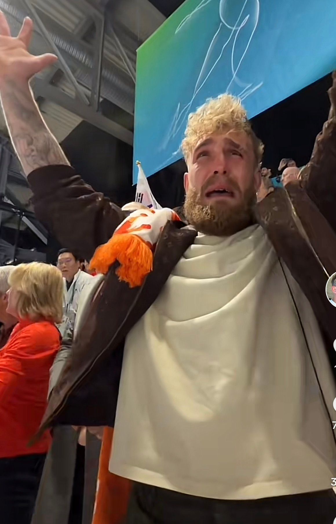 The image captures internet personality and boxer Jake Paul in an exaggerated state of emotional distress, or perhaps mock despair, with tears streaming down his face and his arms dramatically thrown upwards. His curly blonde hair and full beard frame a wide-open mouth, conveying an intense, almost theatrical, reaction. He wears a dark brown jacket over a light-colored shirt, accessorized with a white scarf featuring orange tassels and a small flag, possibly Korean, tucked into his collar. The background suggests an indoor stadium or arena, with a large screen displaying abstract blue and green graphics and glimpses of other spectators. This highly performative display, characteristic of Paul's public persona, adds a humorous layer, as he appears to be reacting with over-the-top emotion to an unseen event, likely a pivotal moment in a sporting contest. The scene perfectly encapsulates the dramatic flair often associated with celebrity reactions at major events.